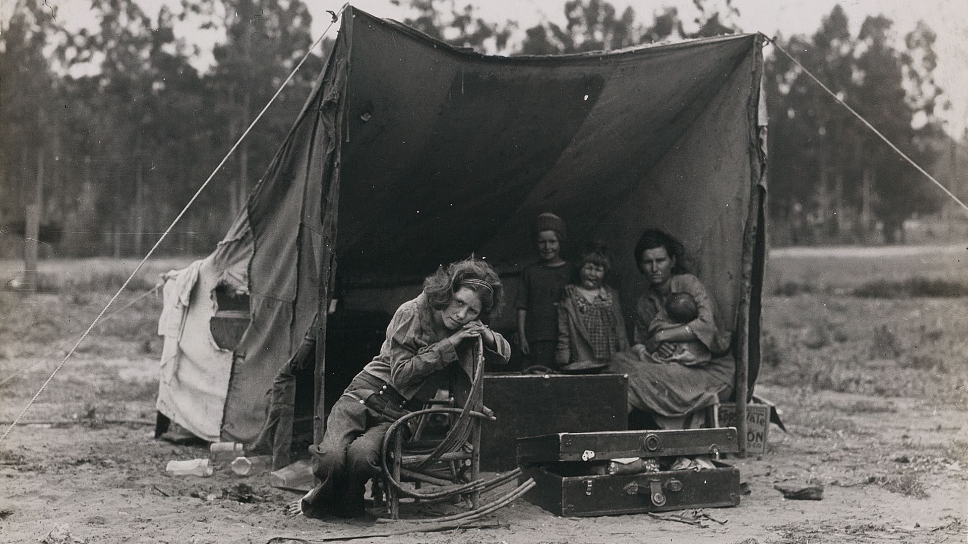 A black-and-white photo of a woman and three children sitting inside a makeshift tent, with one child outside, leaning on a chair; suitcases and belongings are scattered at their feet. Trees are visible in the background.