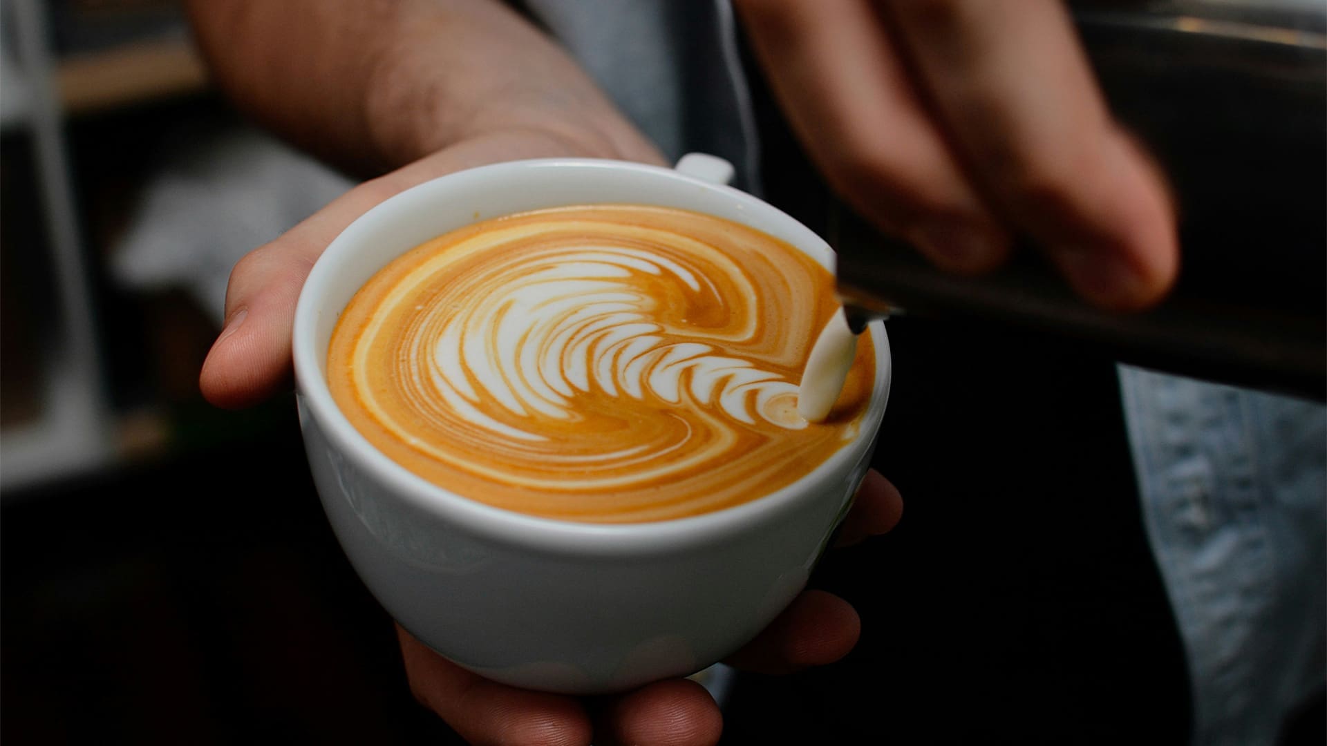 A close-up of a person holding a white cup of coffee with intricate latte art on the surface, as milk is being poured to create a detailed design.