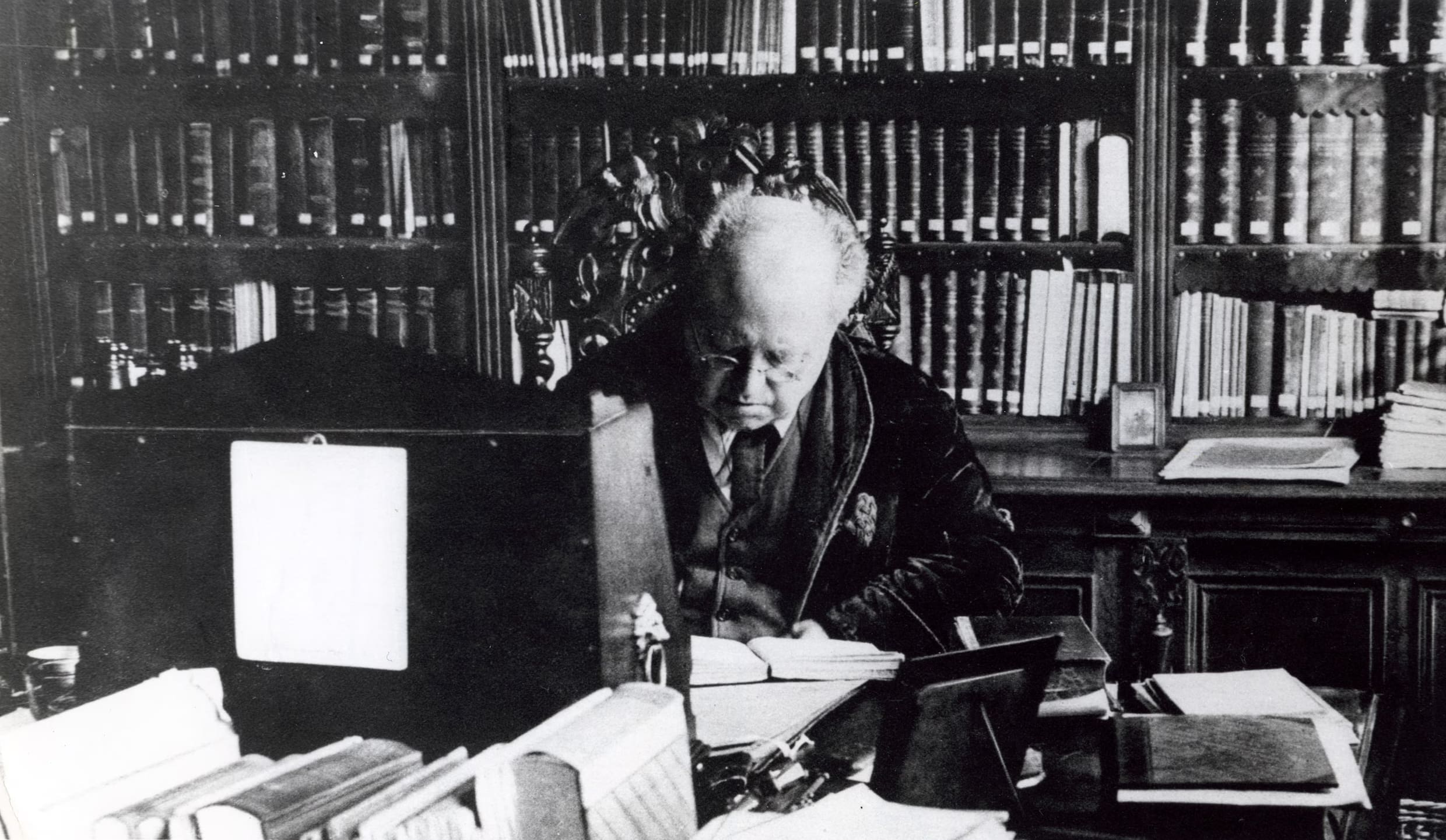 A person with thinning hair sits at a cluttered desk, writing or reading, surrounded by bookshelves filled with books in a dimly lit study or library. The scene appears to be from an earlier historical period.