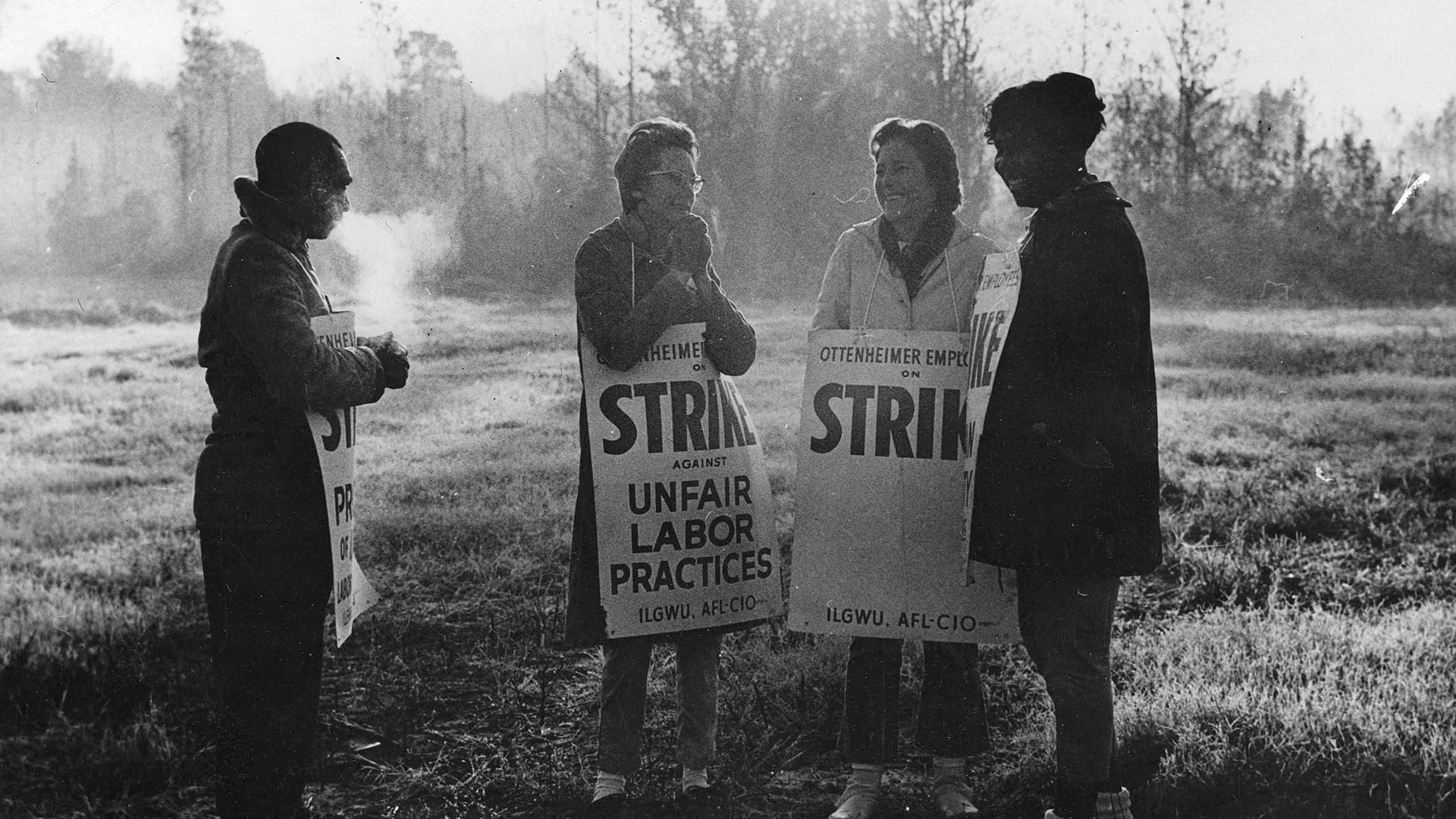 Three female and one male International Ladies Garment Workers Union members stand in the cold holding strike placards protesting unfair labor practices by the Ottenheimer company.