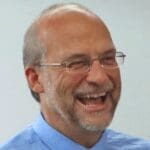 A woman with dark hair pulled back, wearing a dark top and a green cardigan, smiles at the camera in a softly lit indoor setting.