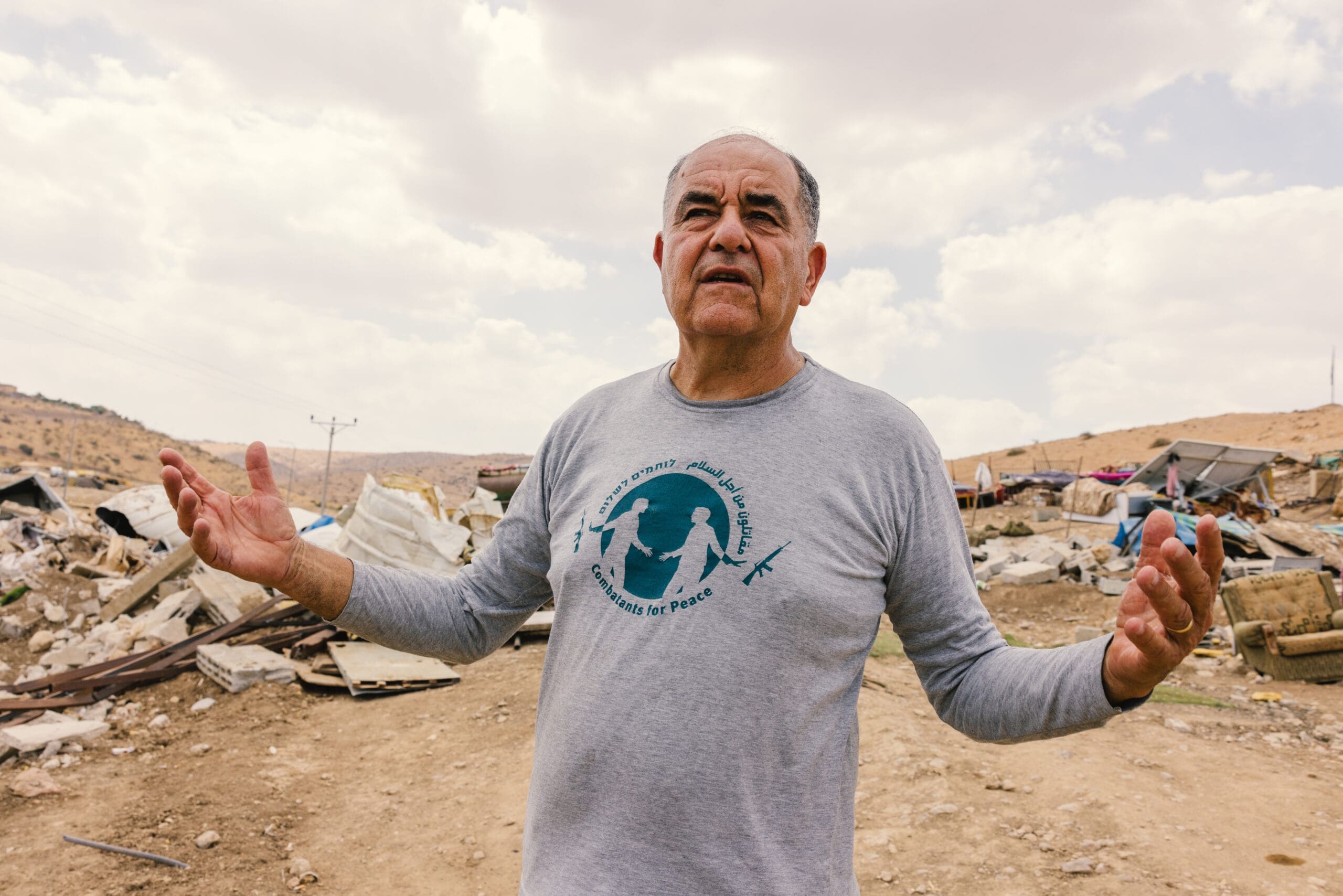 An older man stands outdoors with his arms raised, wearing a gray long-sleeve shirt with a dove and text. Behind him are scattered debris and damaged structures under a cloudy sky in a rural, hilly area.