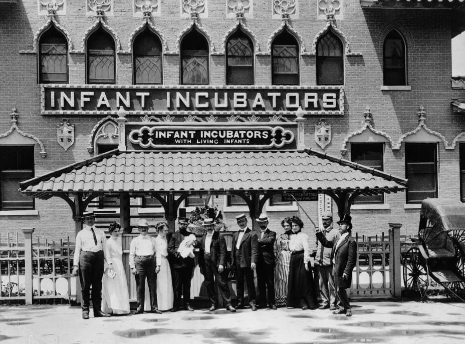 A group of people stand in front of an ornate brick building with a sign reading Infant Incubators with Living Infants, likely from the early 1900s. Women in long dresses and men in suits and hats pose by a decorative fence.