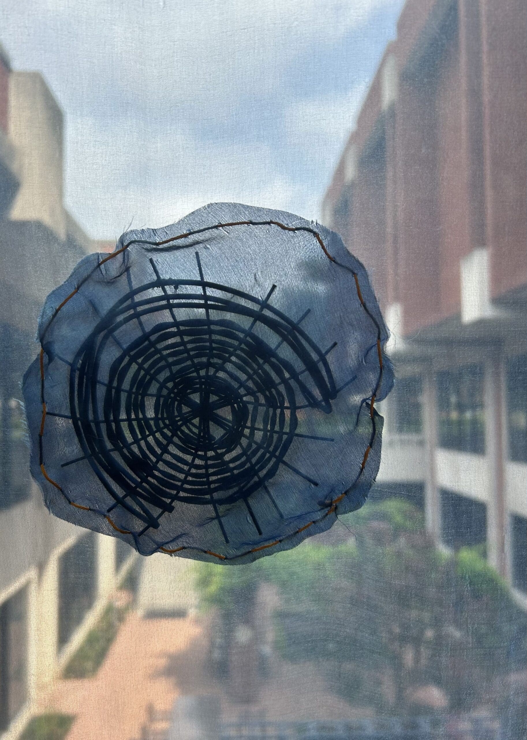 A circular piece of translucent fabric with black geometric stitching is attached to a window, overlooking a courtyard and tall buildings under a partly cloudy sky.