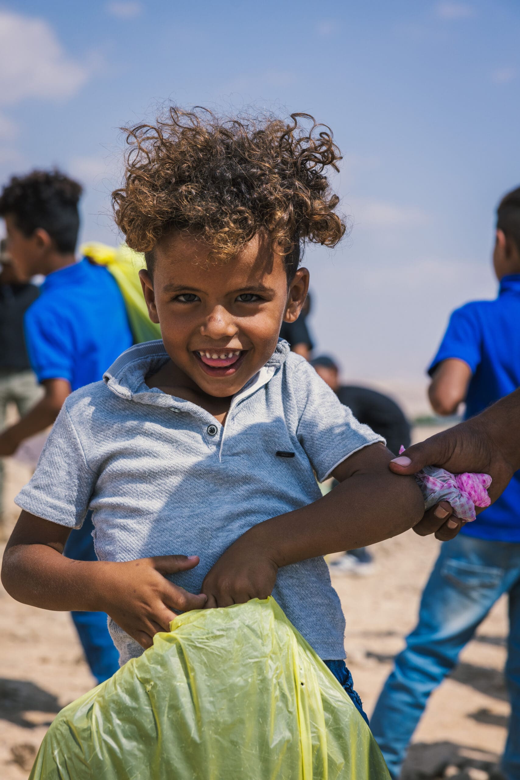 A young child with curly hair, wearing a gray shirt, smiles at the camera while holding a yellow bag. Other children in blue shirts stand in the background outdoors under a blue sky.