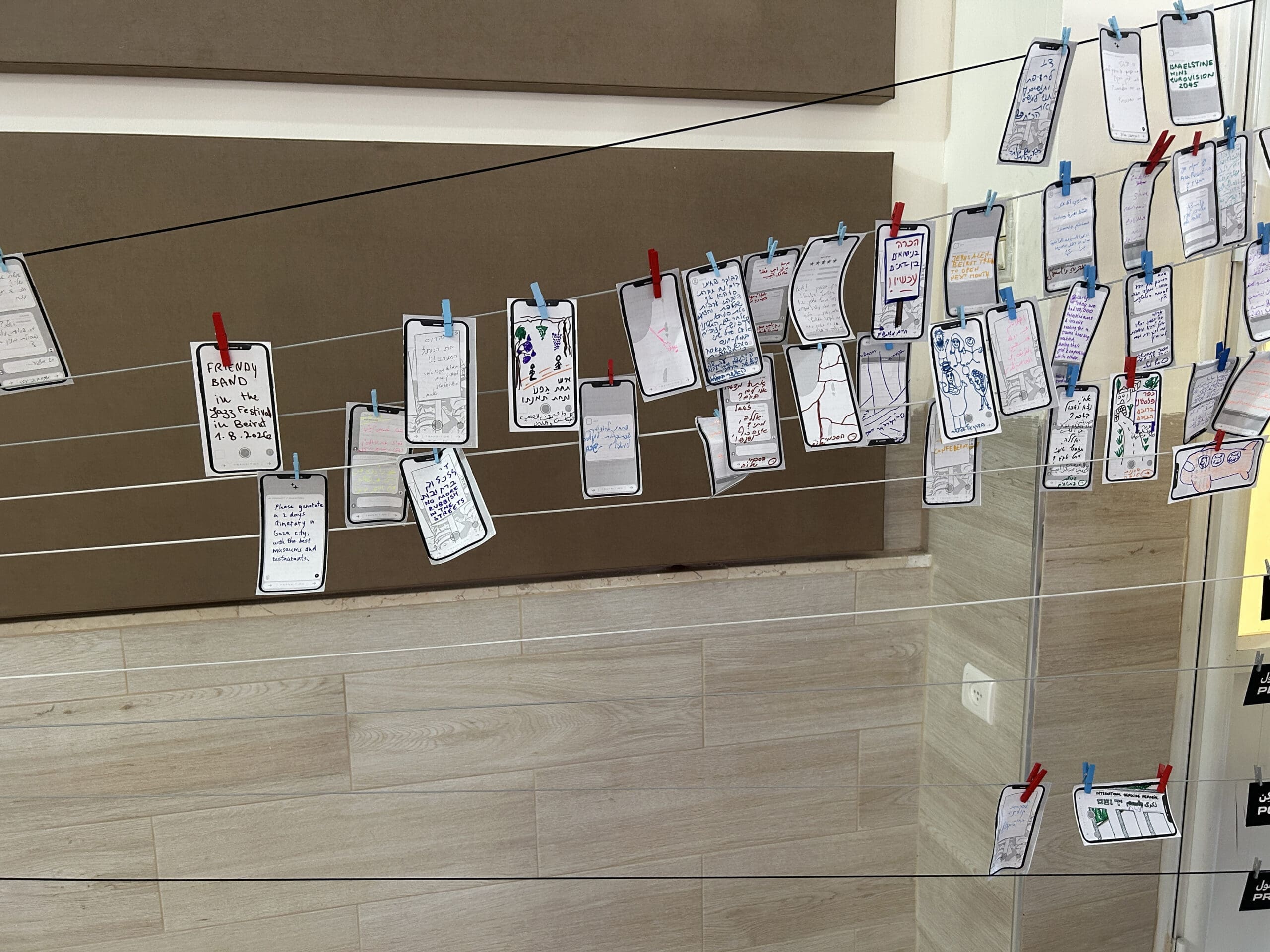 Small illustrated papers resembling smartphone screens are clipped to strings with colorful clothespins, hanging against a beige wall and wooden floor. The papers display handwritten notes, drawings, and colorful decorations.