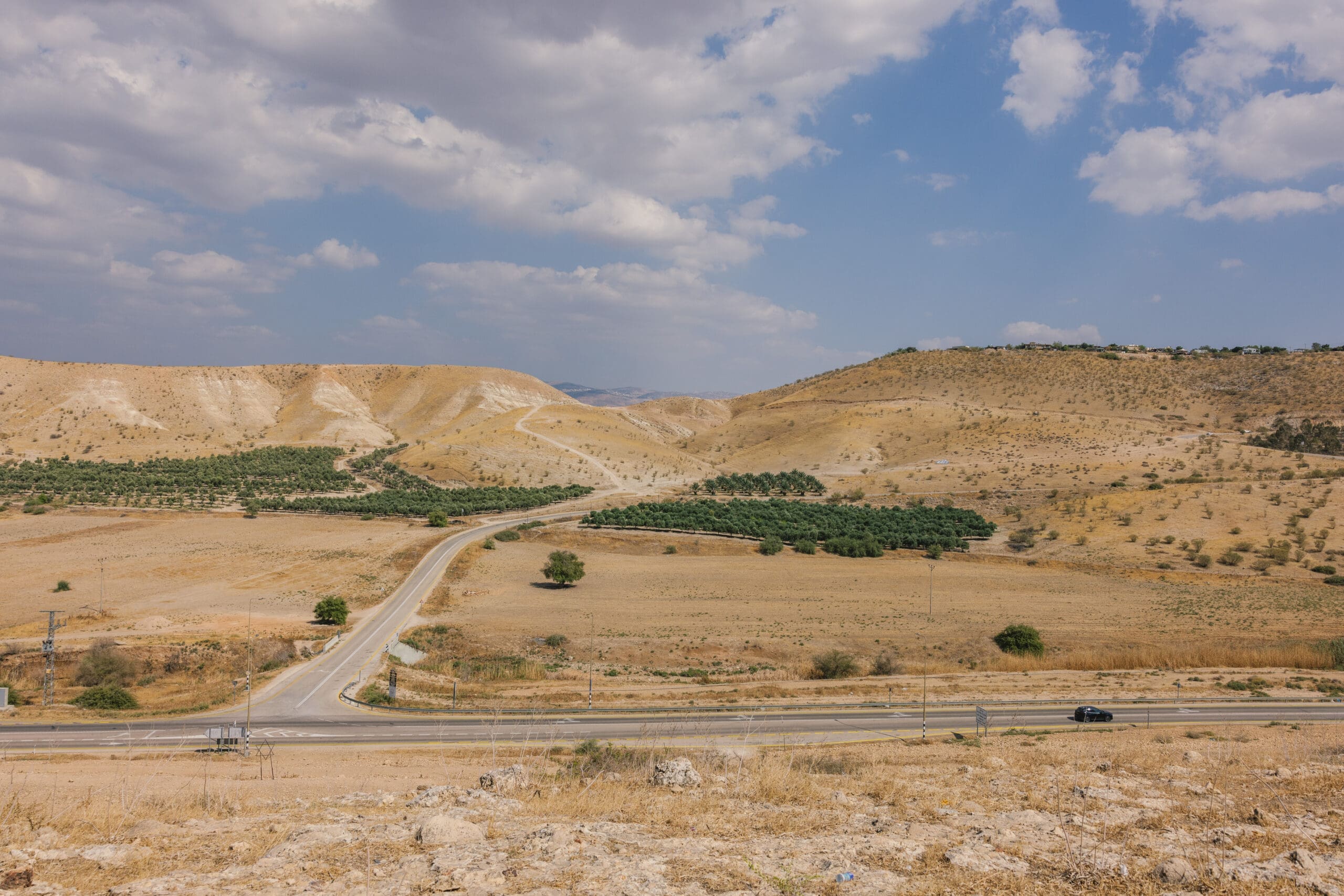 A winding road runs through a dry, hilly landscape with patches of green trees and shrubs under a partly cloudy sky. A lone car is visible on the road in the foreground.