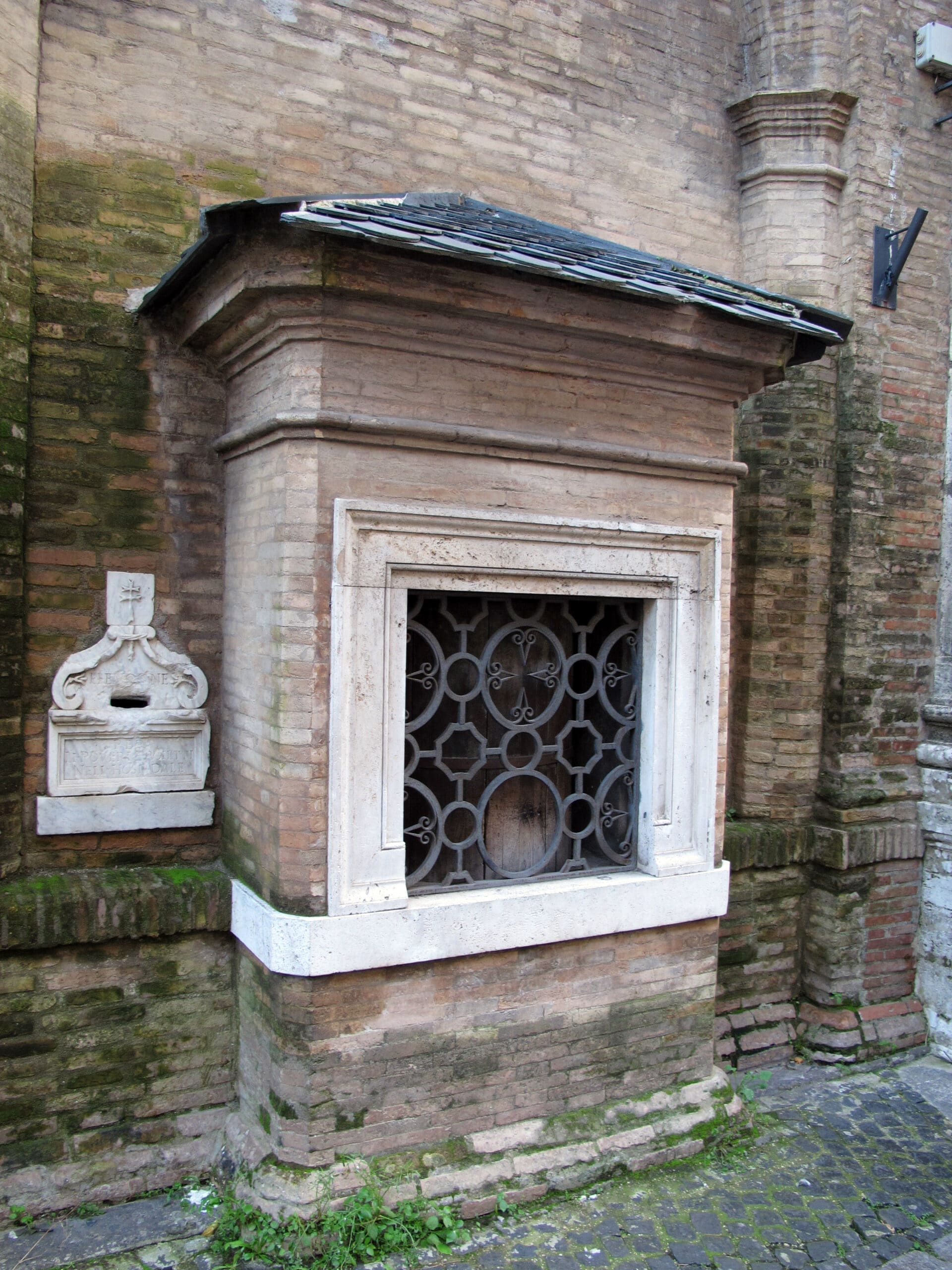 A stone structure with a decorative iron grate window set in a brick wall, next to a small stone plaque with a slot, possibly used for donations or messages. Moss grows on the ground below.