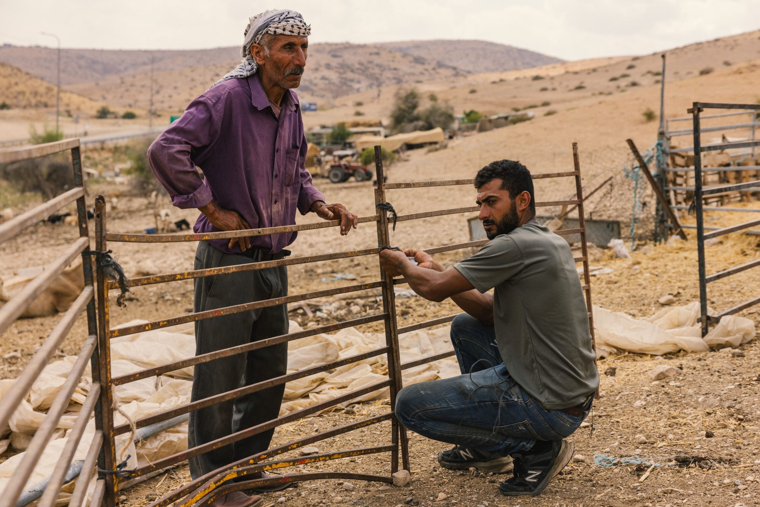Two men work together on a metal fence in a dry, rural landscape. One stands holding the fence, while the other kneels, attaching or repairing part of it. Hills and scattered equipment are visible in the background.