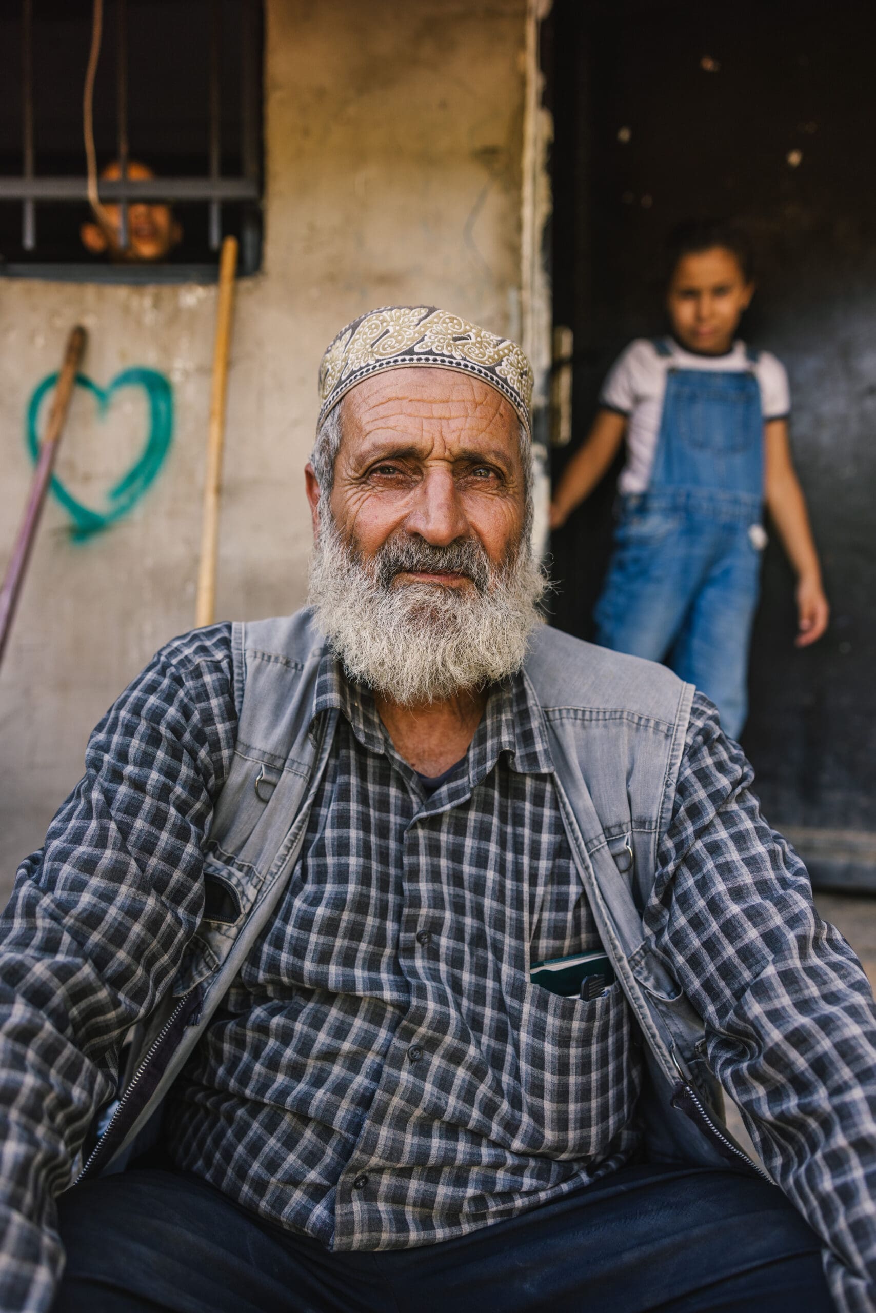 An elderly man with a gray beard and patterned cap sits outdoors, wearing a plaid shirt and vest. Behind him, a child in overalls stands near a dark doorway, and another child looks out from a barred window. A green heart is painted on the wall.