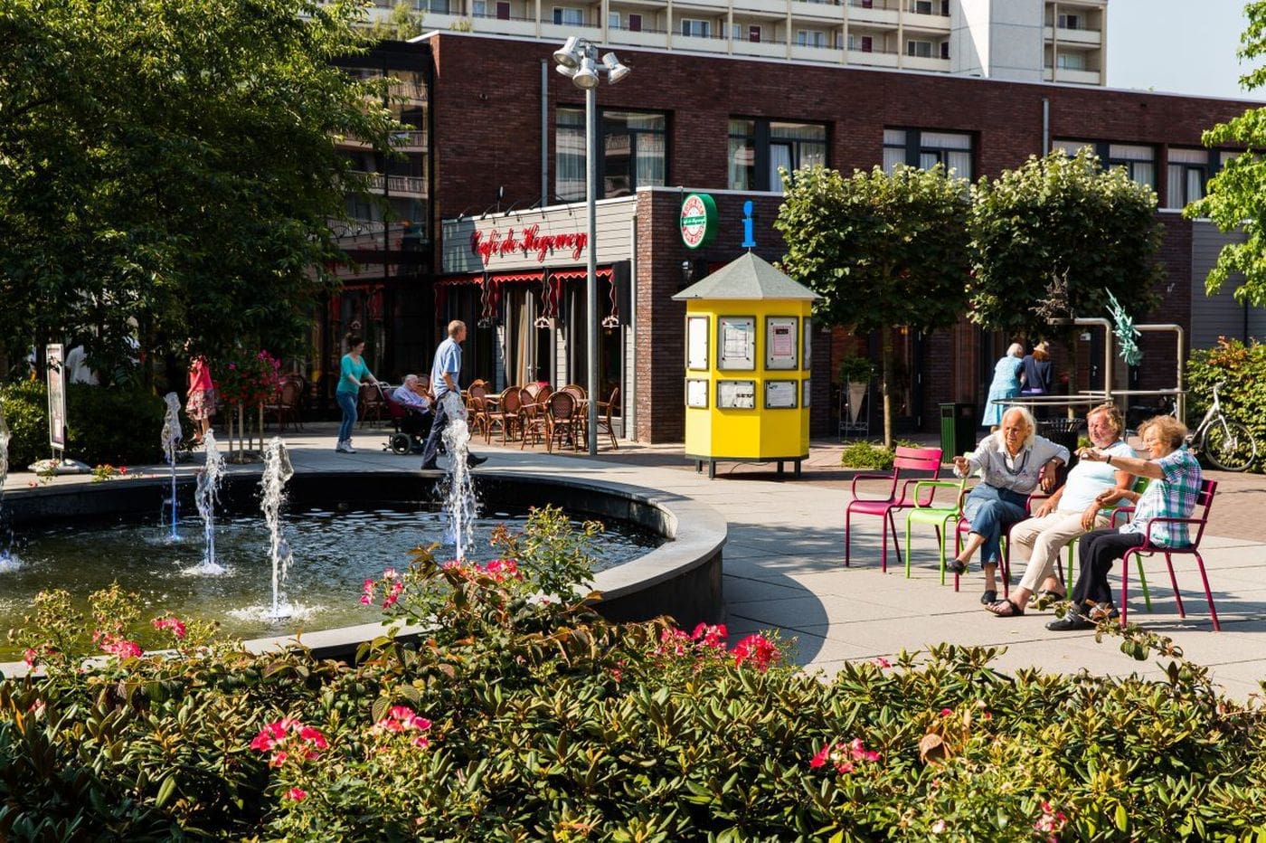 People relax on colorful chairs near a circular fountain with water jets in a sunny plaza. Pink flowers and bushes surround the area, with buildings, a cafe, and a yellow kiosk in the background.