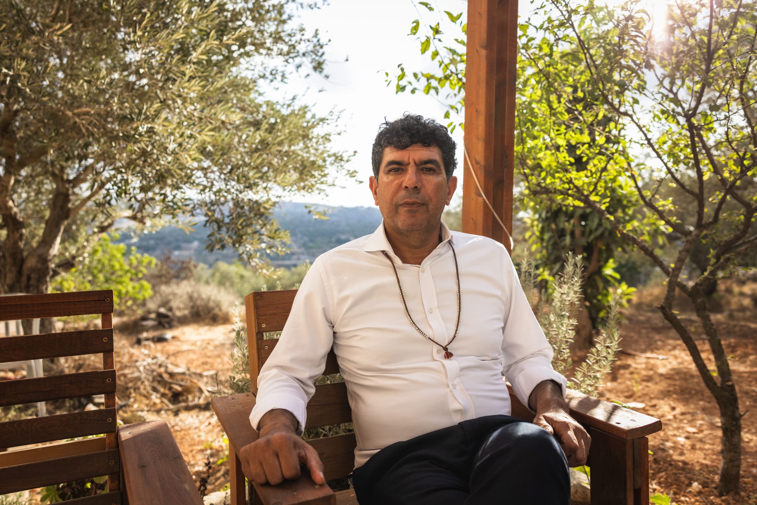 A man in a white shirt sits on a wooden chair outdoors, surrounded by trees and greenery, with sunlight filtering through the leaves. He looks calmly at the camera, hands resting on the chair arms.