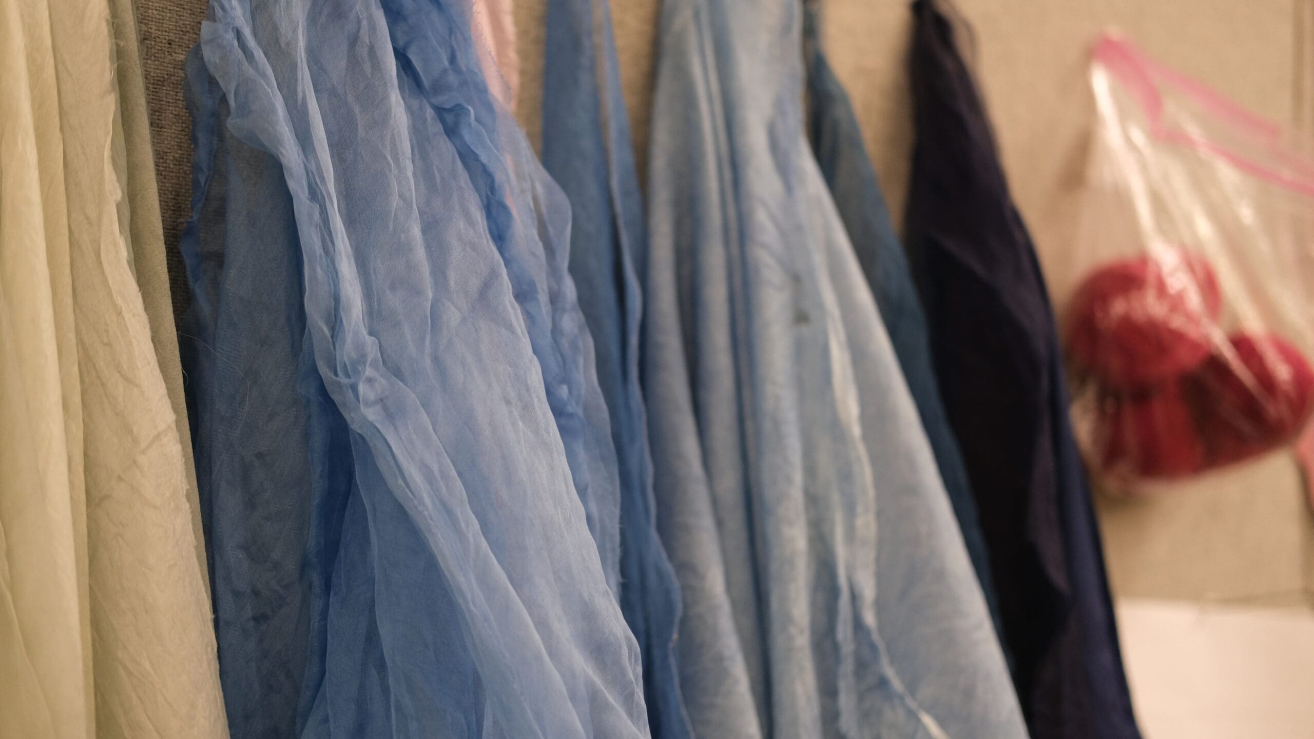Sheer fabric scarves in various shades of blue and cream hang side by side, with a plastic bag containing red yarn visible in the background.