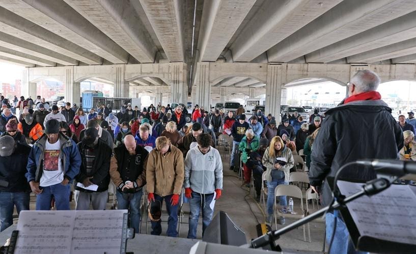A large group of people stands and sits under a highway overpass, many with heads bowed, during a gathering or event. A person speaks from a podium in the foreground, with musical equipment visible nearby.