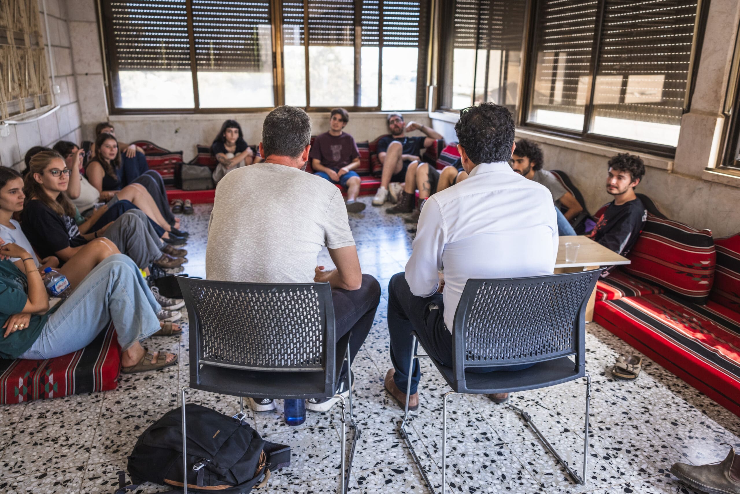 A group of people sit in a semi-circle on cushions and chairs in a sunlit room, listening to two men at the front who are leading a discussion. The atmosphere appears casual and focused.