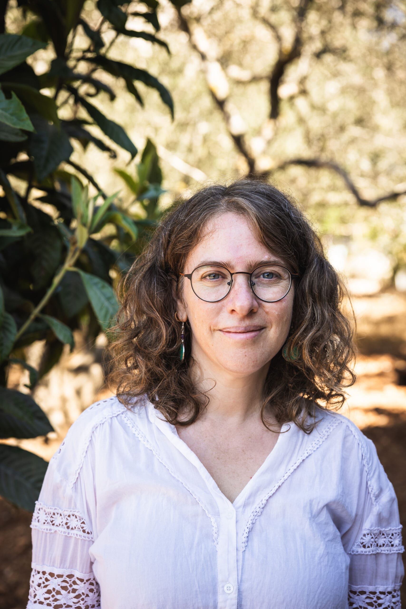 A woman with curly brown hair and glasses, wearing a white blouse, stands outdoors near greenery with sunlight filtering through the trees in the background. She is smiling gently at the camera.