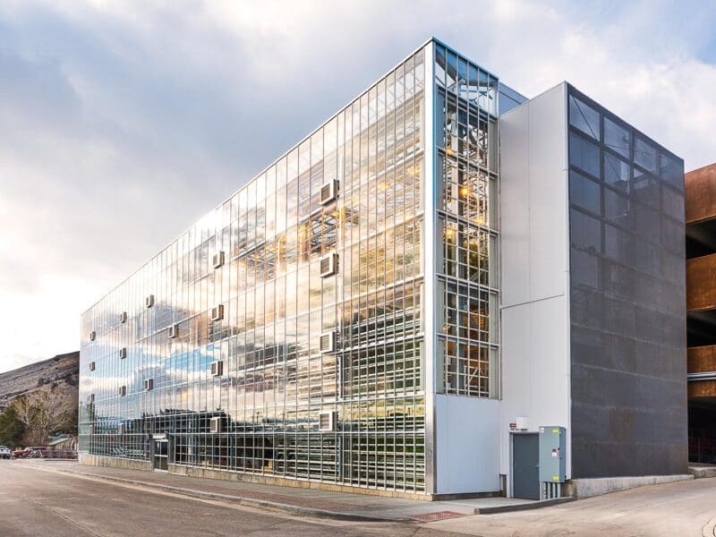 A modern multi-story building with a reflective glass facade, showing sky and clouds, and a metallic structure on one side. The scene appears industrial and is situated near a mountain.