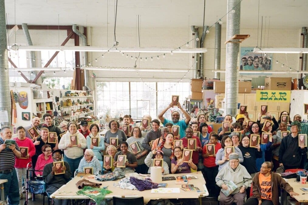 A large group of people in an art studio, many holding up portraits. The room is filled with art supplies, colorful decorations, and tables, with natural light streaming through big windows.