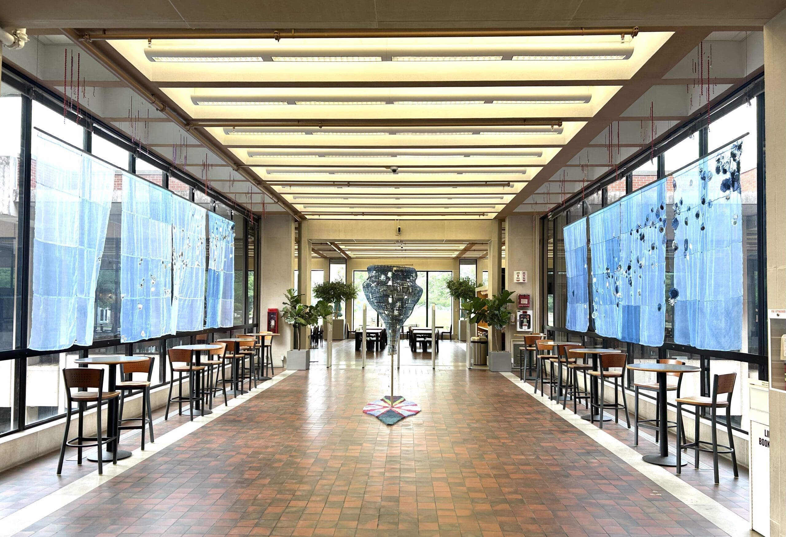 A spacious, modern hallway with tall windows covered in blue art panels. Small tables and chairs line both sides, and plants are arranged at the far end near glass doors. The floor is tiled in shades of brown.