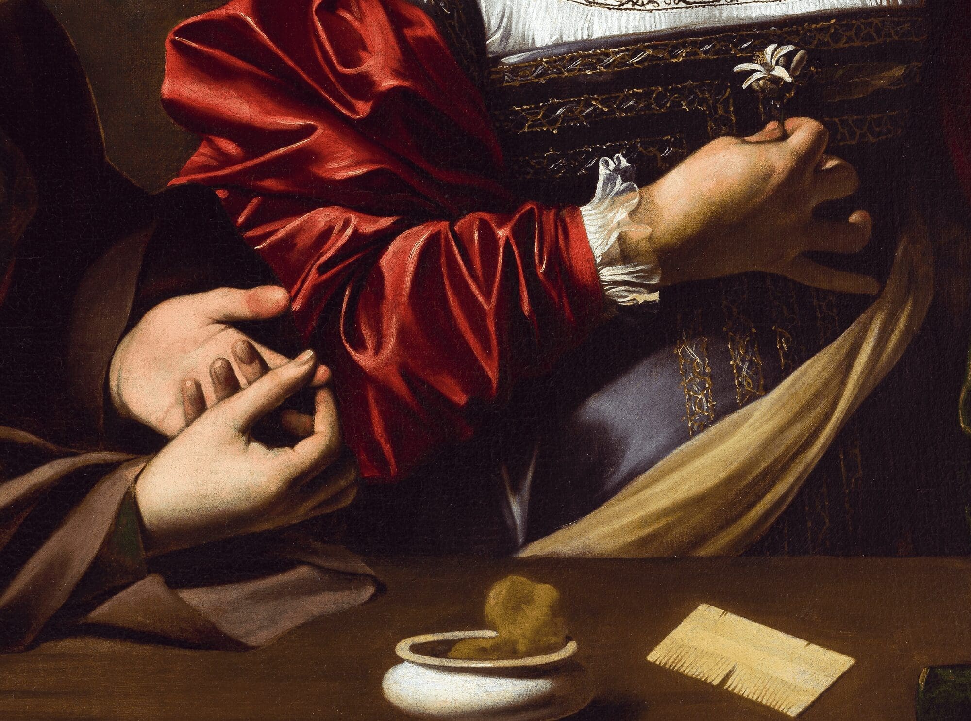 A close-up of two people’s hands: one person in a red and white dress holds a white lily, while another gently holds their arm. Objects, including a comb and a white ring, rest on a nearby table.