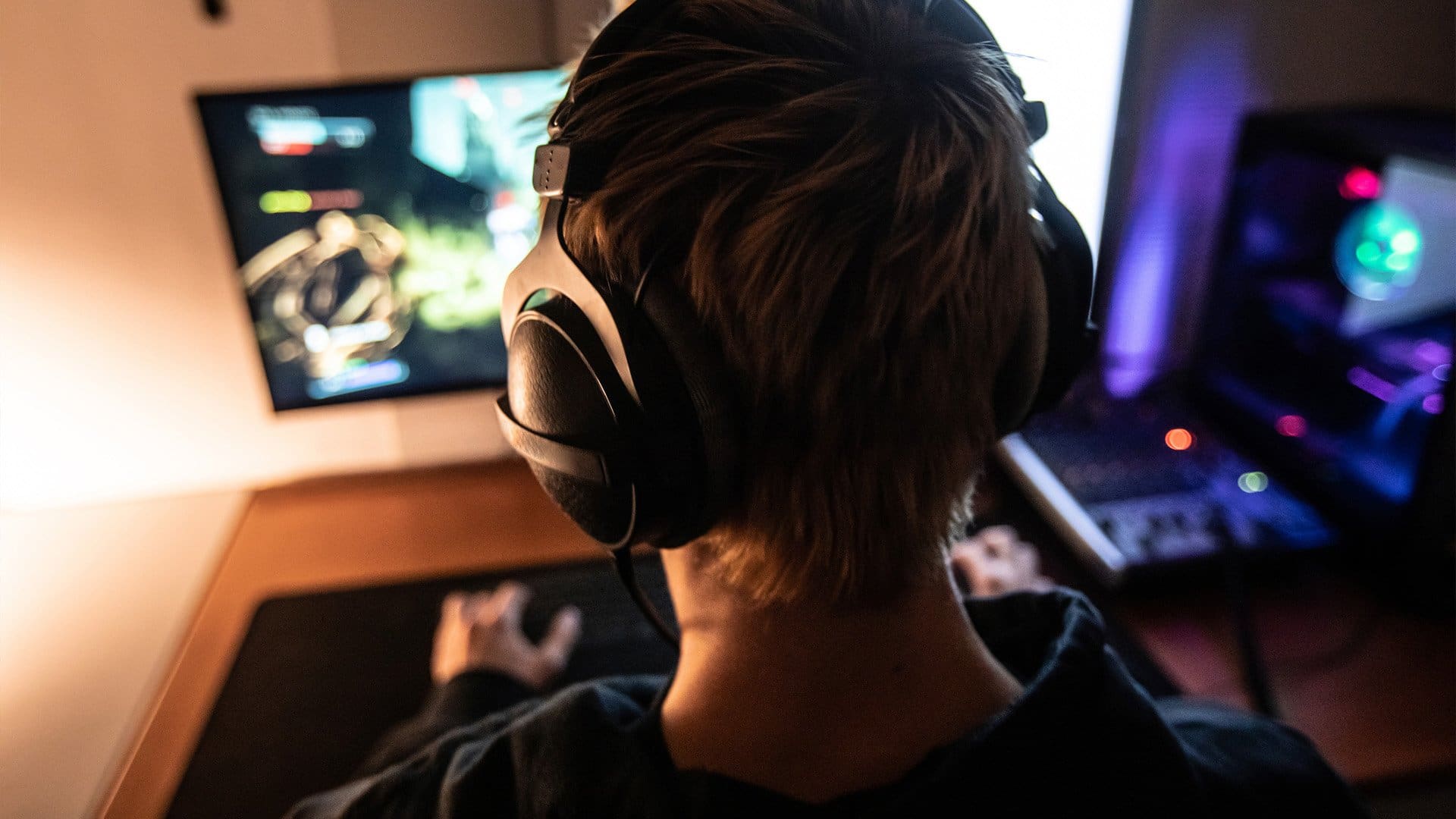 A person wearing large headphones sits at a desk, playing a video game on a computer monitor in a dimly lit room with a gaming PC and colorful lights in the background.
