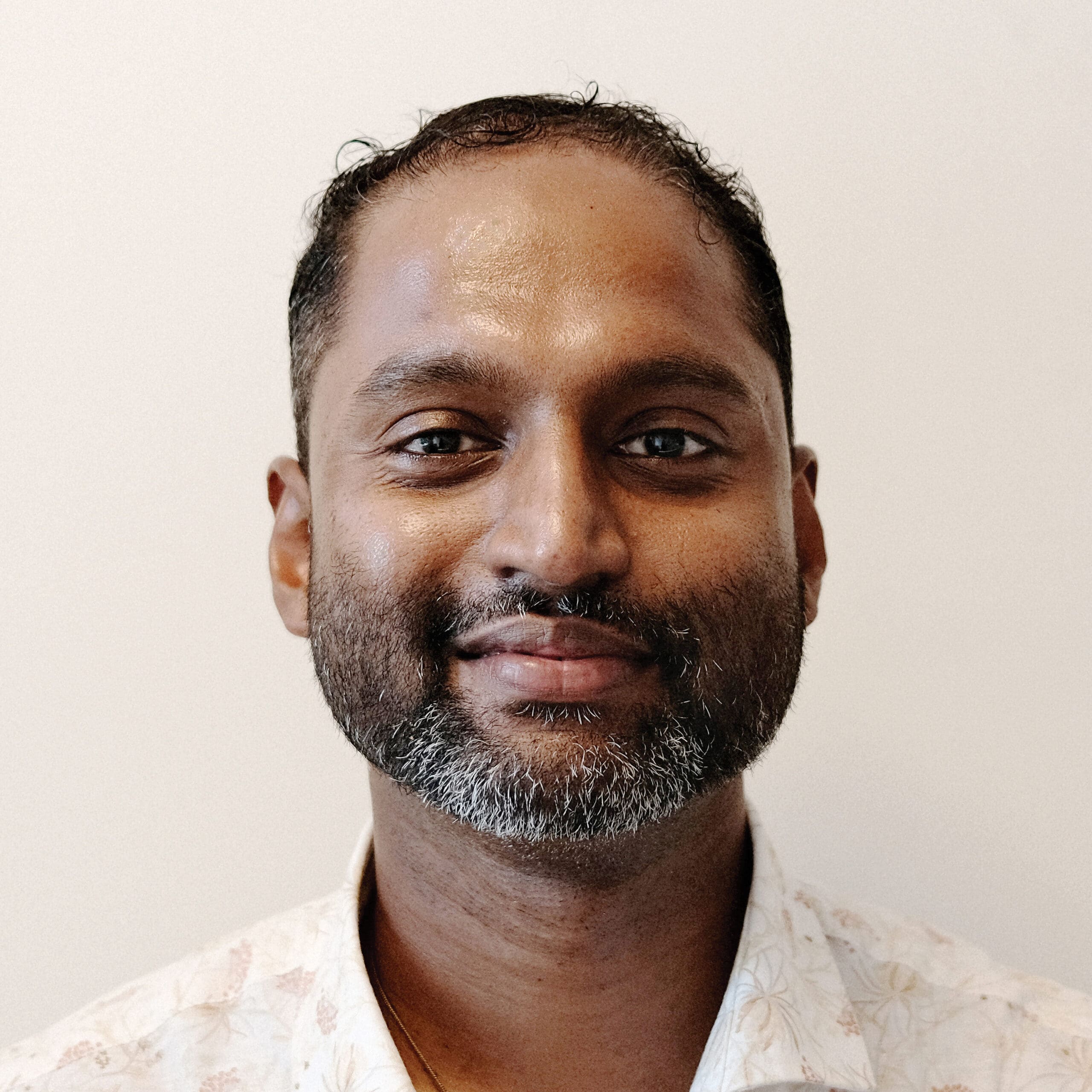 A man with short dark hair and beard, wearing a light-colored collared shirt, is smiling gently in front of a plain light background.