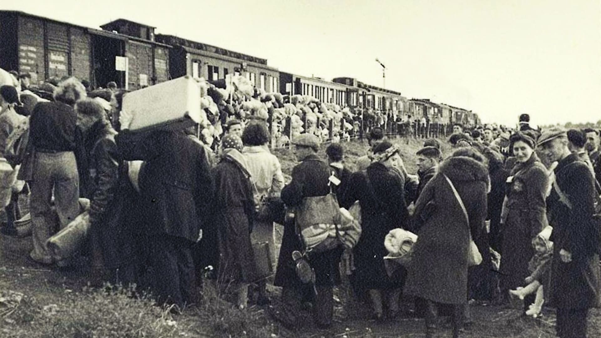 A large group of people carrying bags and belongings gathers near a train with open boxcars, preparing to board under overcast skies. The scene appears historical, with people dressed in early-to-mid 20th century clothing.