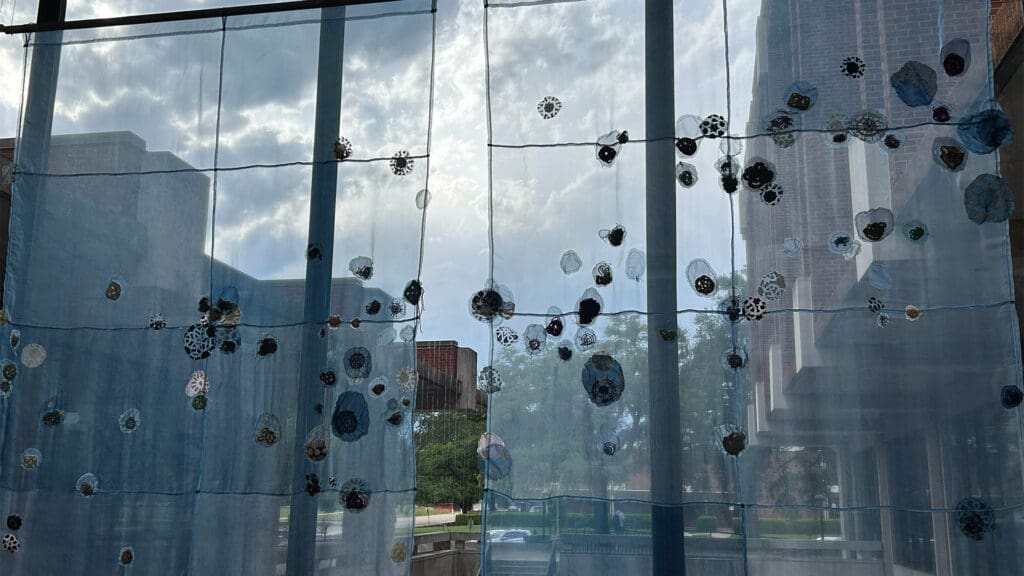 A sheer curtain decorated with fabric circles hangs in front of tall windows. Outside, brick buildings, trees, and a cloudy sky are visible through the glass.
