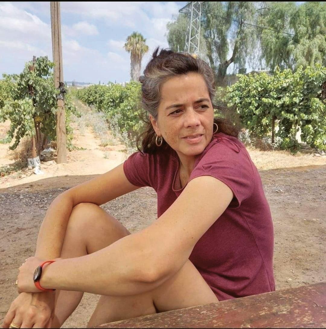 A woman with brown hair and a streak of gray, wearing a maroon shirt and shorts, sits outdoors on a sunny day near green grapevines, looking thoughtful. Trees and vineyard rows are in the background.
