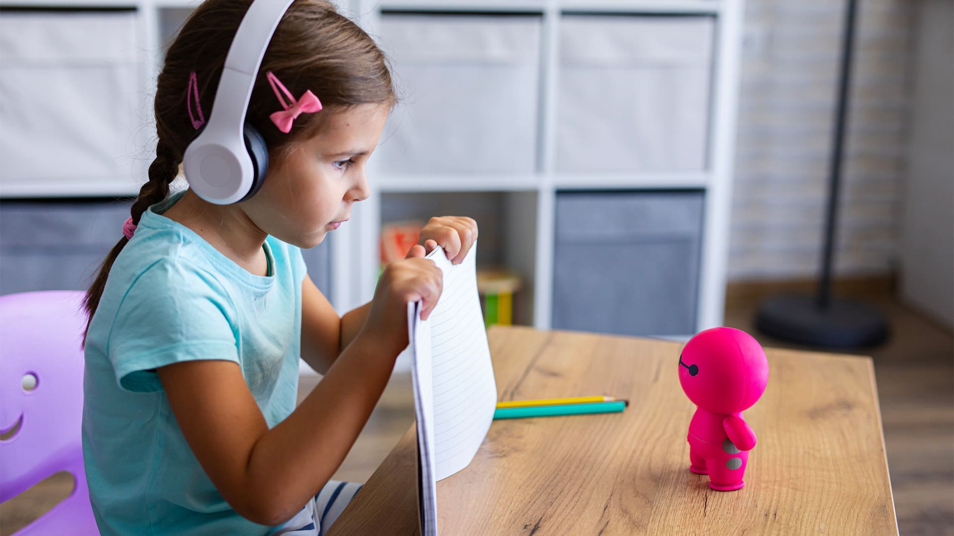 A young girl wearing headphones and holding a notebook sits at a desk, facing a small pink robot toy. A pencil and eraser are on the desk, and shelves are visible in the background.