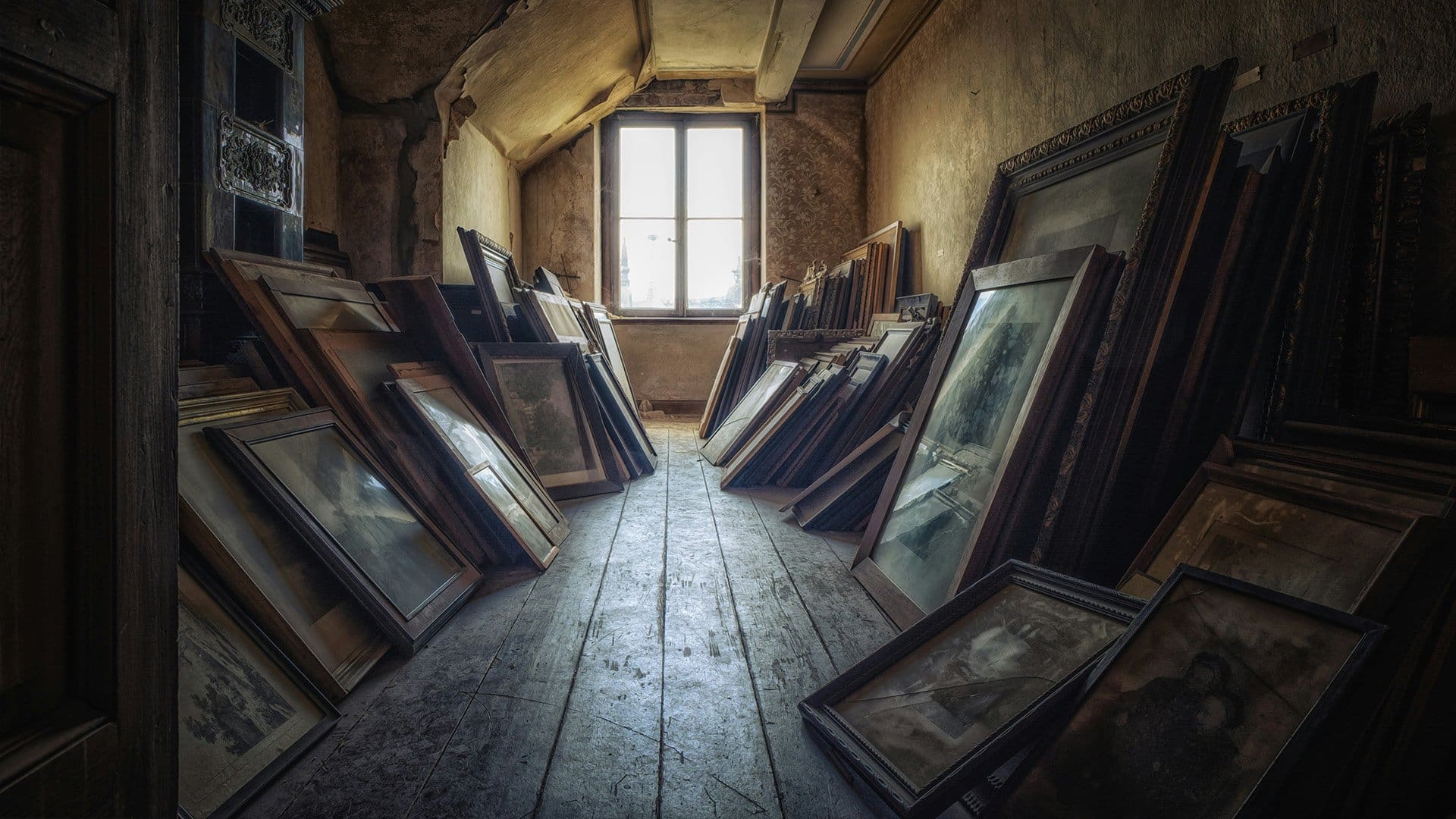 Dusty, old picture frames and paintings are stacked against the slanted walls of a dimly lit attic room with wooden floors and a single window letting in light.