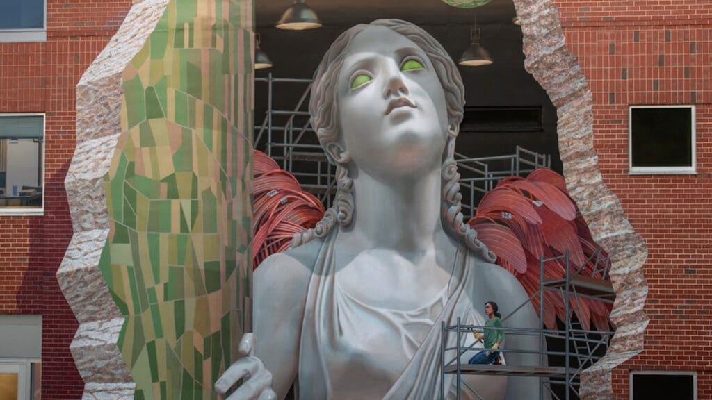 A person stands on scaffolding beside a large statue of a woman with glowing green eyes, emerging from a brick wall as if breaking through it. The statue is surrounded by vibrant, colorful shapes and detailed textures.