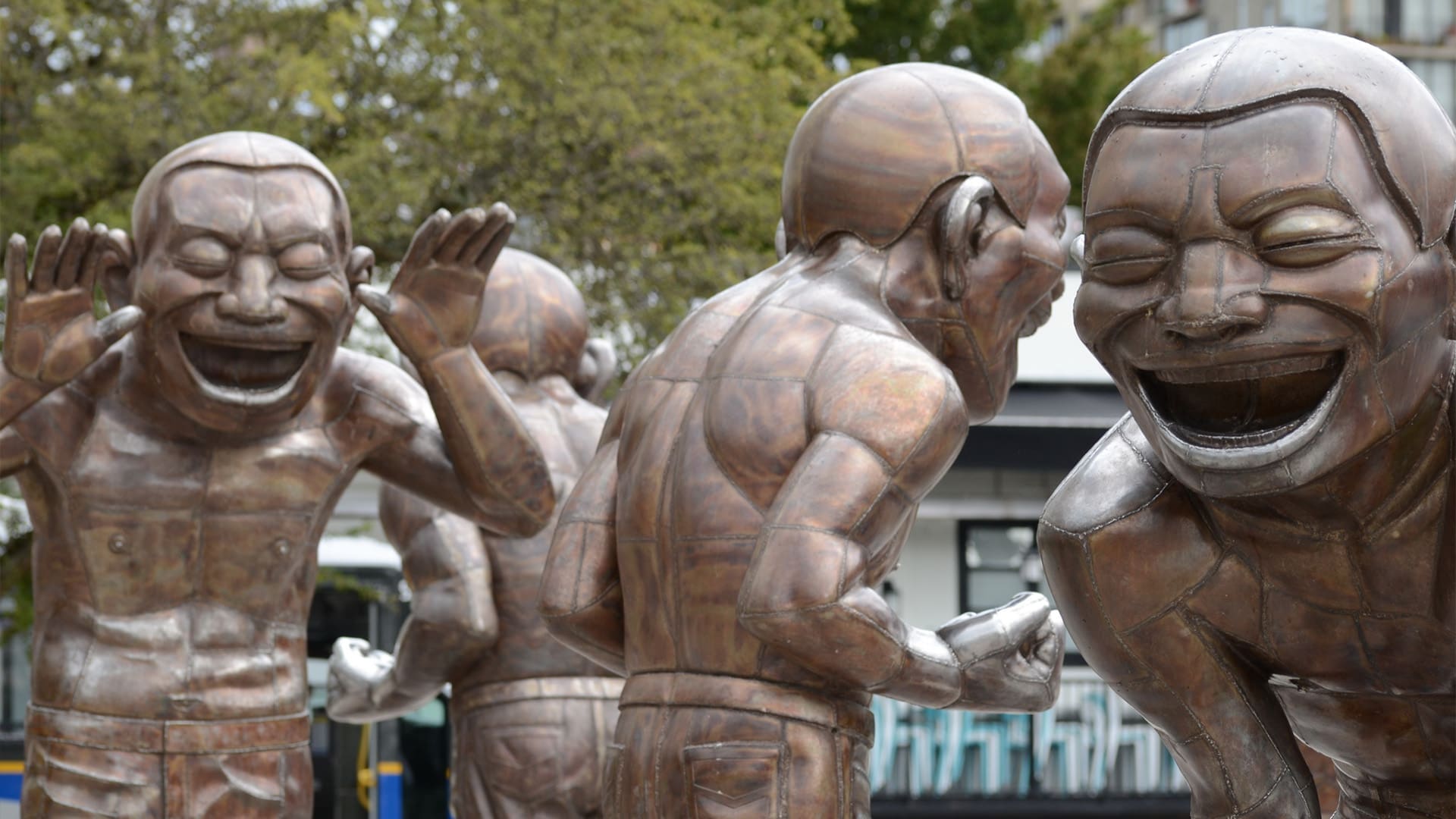 Bronze statues of muscular figures laughing with exaggerated facial expressions, standing in various playful poses outdoors, with trees and buildings in the background.