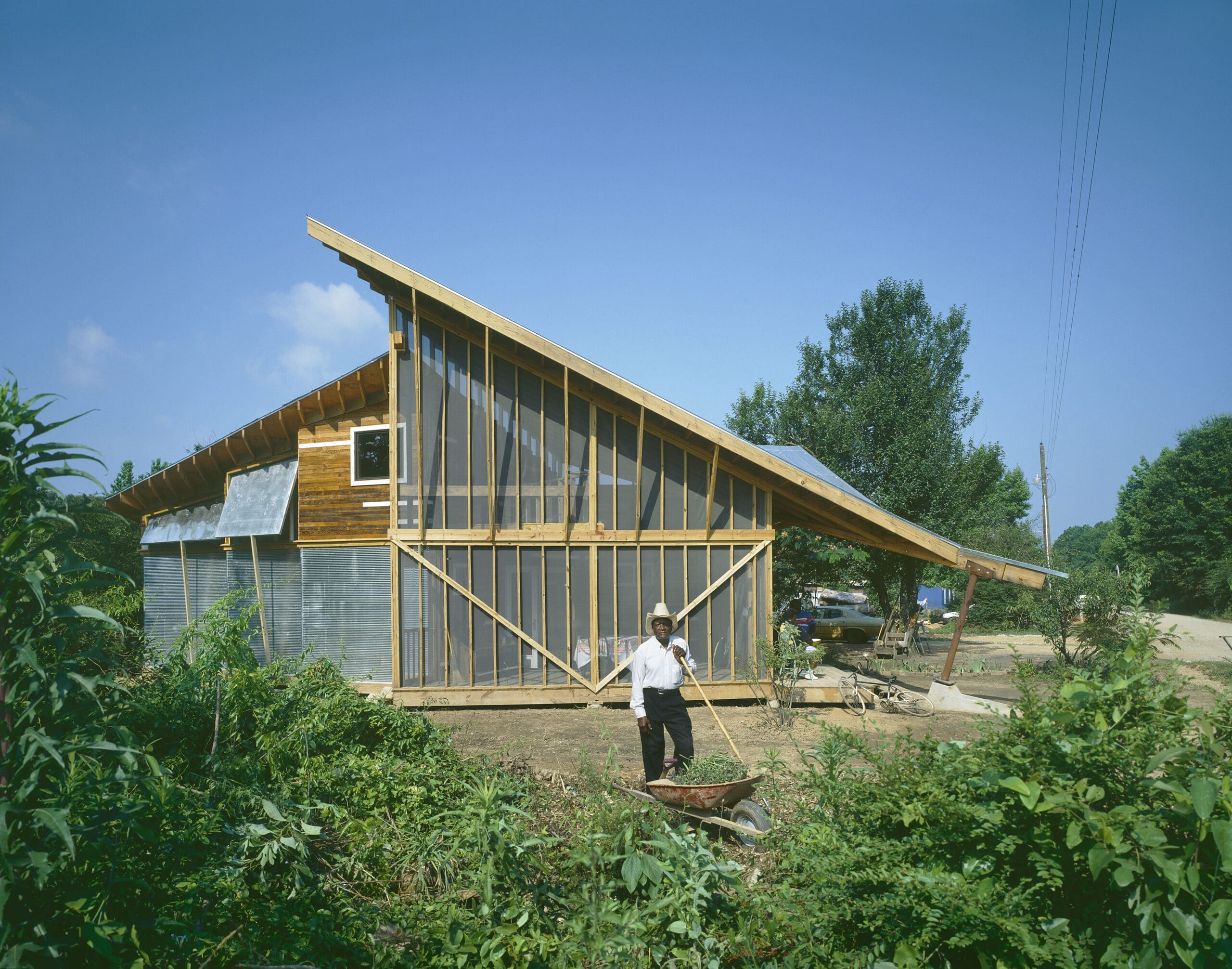 A person with a hat pushes a wheelbarrow in front of a modern house with a steep slanted roof, wood and metal siding, large windows, and a lush green garden under a clear blue sky.