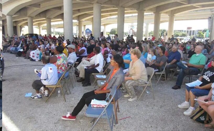 A large, diverse group of people sits on folding chairs under a highway overpass, attentively facing forward as though attending an outdoor event or gathering. Daylight filters through the columns overhead.