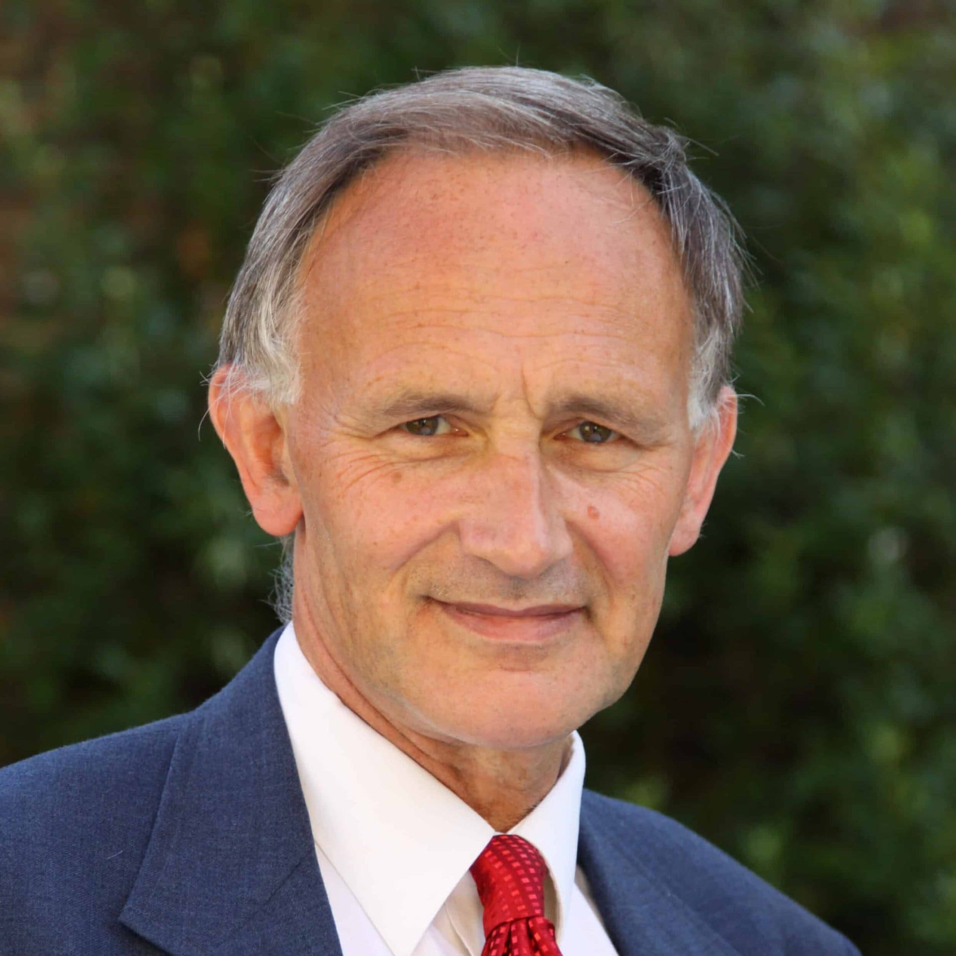 A middle-aged man with short gray hair, wearing a blue suit, white shirt, and red tie, smiles gently while standing outdoors in front of green foliage.