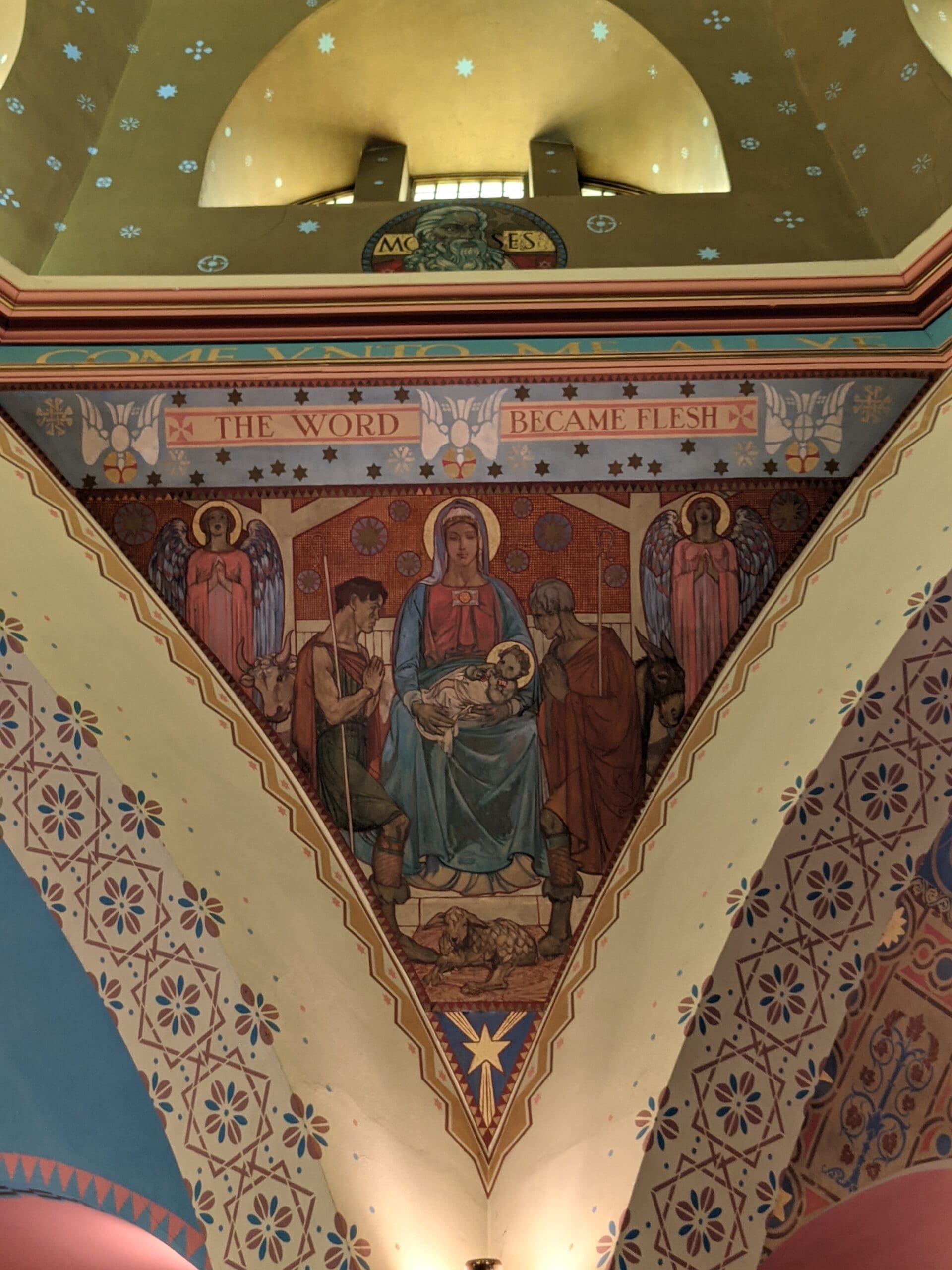 A painted church mural shows Mary holding the infant Jesus, surrounded by angels and figures. Above, a banner reads THE WORD BECAME FLESH. The scene is ornate with vibrant colors and decorative patterns.