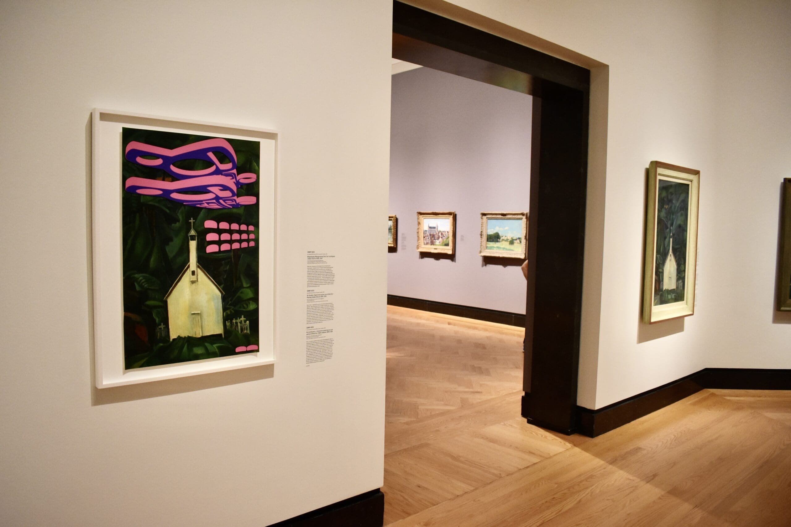 A museum gallery with framed paintings on white walls; one artwork in the foreground shows a small house with pink abstract shapes above it, while more landscape paintings hang in the adjoining room.