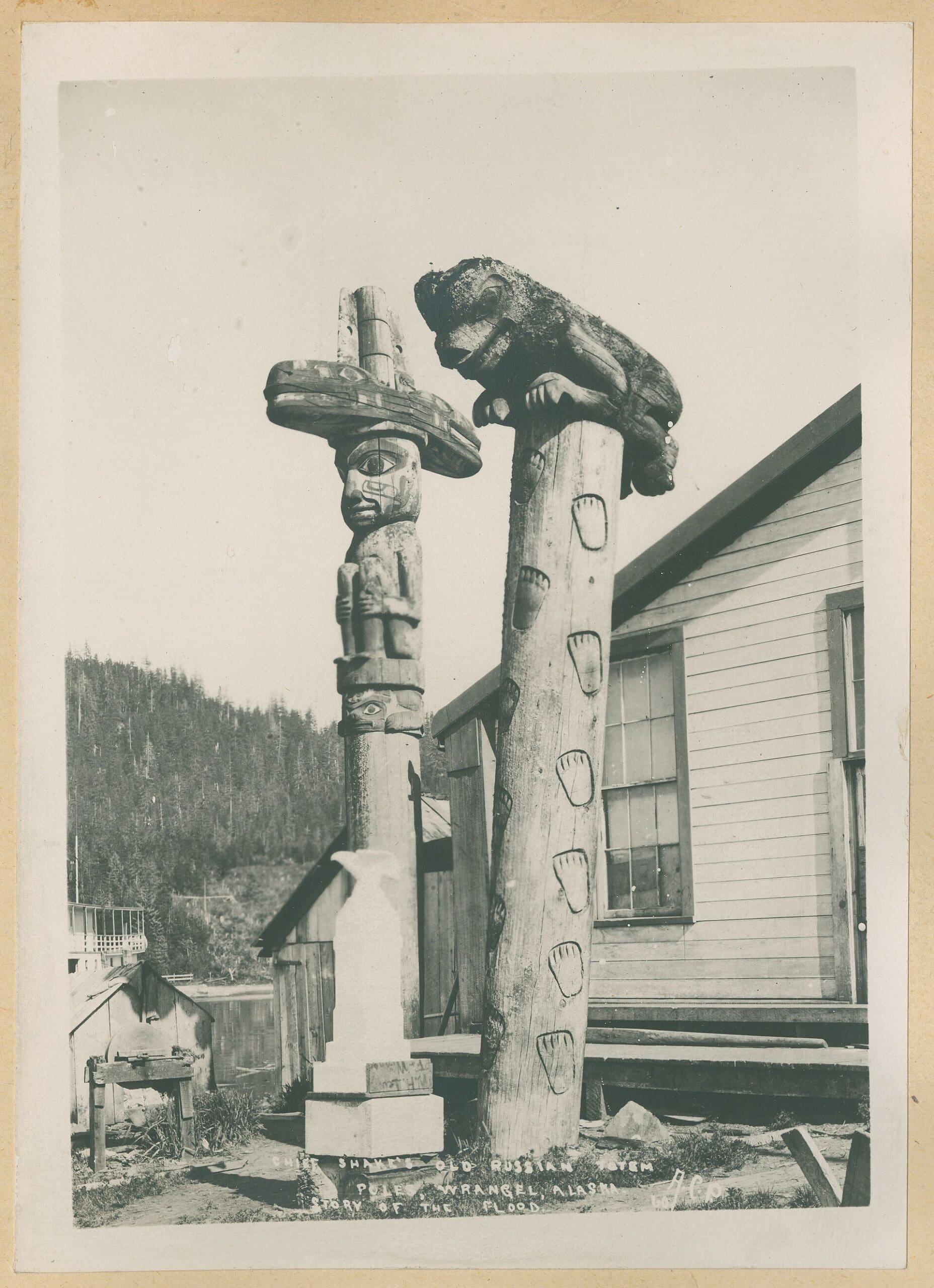 Two carved totem poles stand outside wooden buildings; one features a human figure, and the other is topped with a bear. Forested hills are visible in the background.