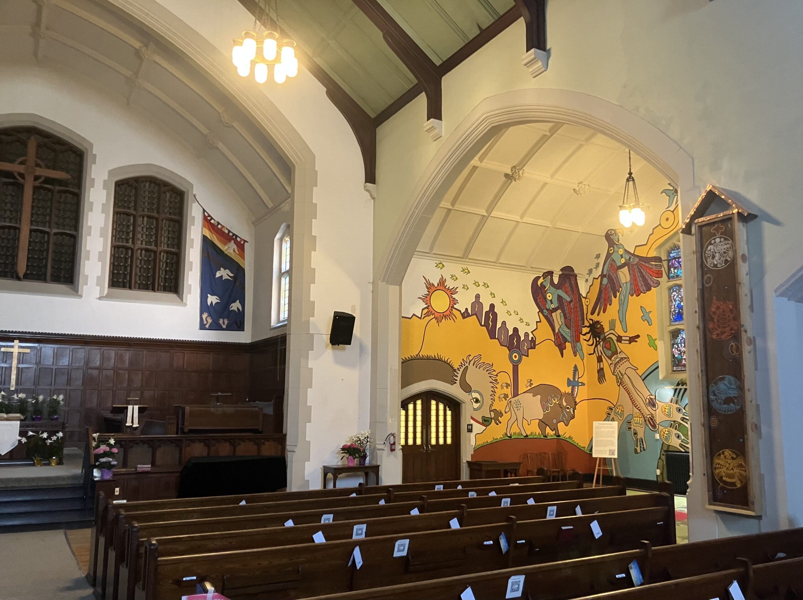 Interior of a church with wooden pews, stained glass windows, and altars. One wall features a colorful mural with animals and abstract shapes, while the rest of the room is more traditional in style. Ceiling lights hang overhead.