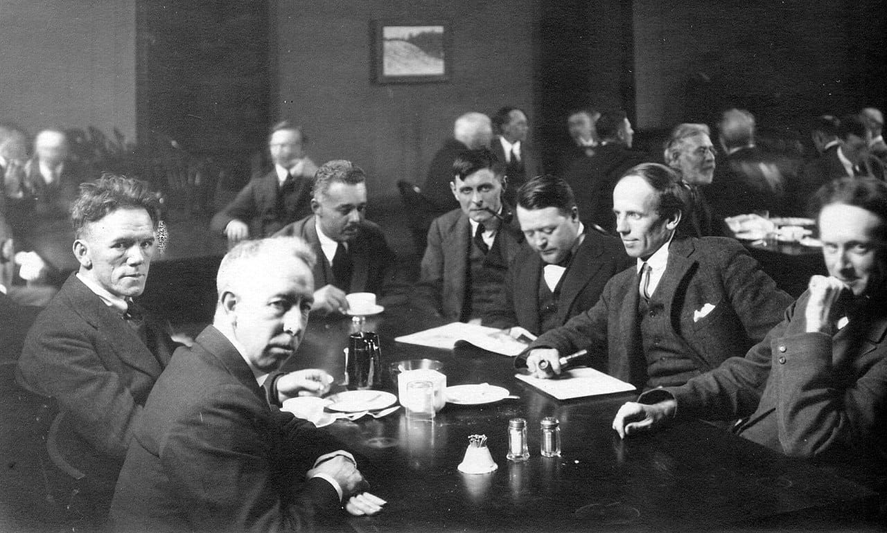 A group of men in suits sit around a table with papers, cups, and a coffee pot in a formal setting, while others converse in the background. The scene appears to be from the early 20th century.