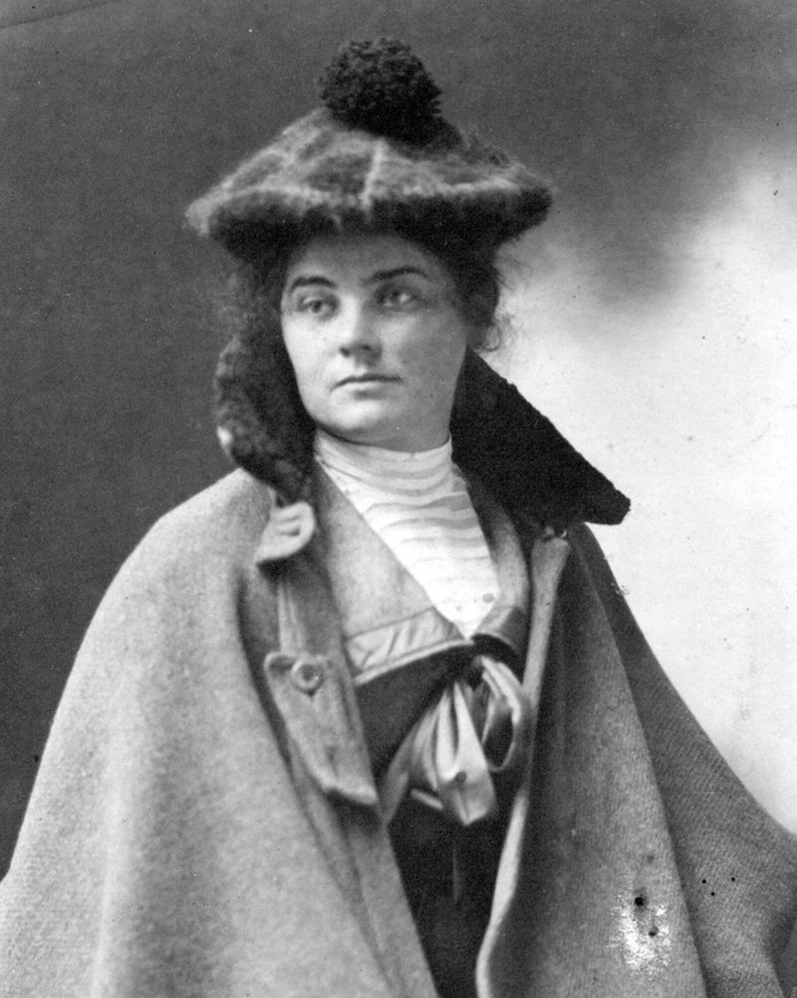 Black-and-white portrait of a woman wearing a textured, wide-brimmed hat with a pom-pom, a high-collared blouse, and a cape-style coat. She gazes slightly to the side with a neutral expression.