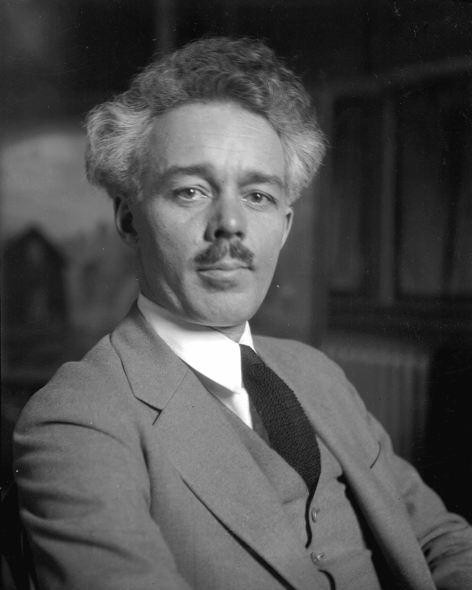 Black and white portrait of a man with wavy gray hair and a mustache, wearing a suit, tie, and vest, looking directly at the camera with a neutral expression. The background is softly blurred.