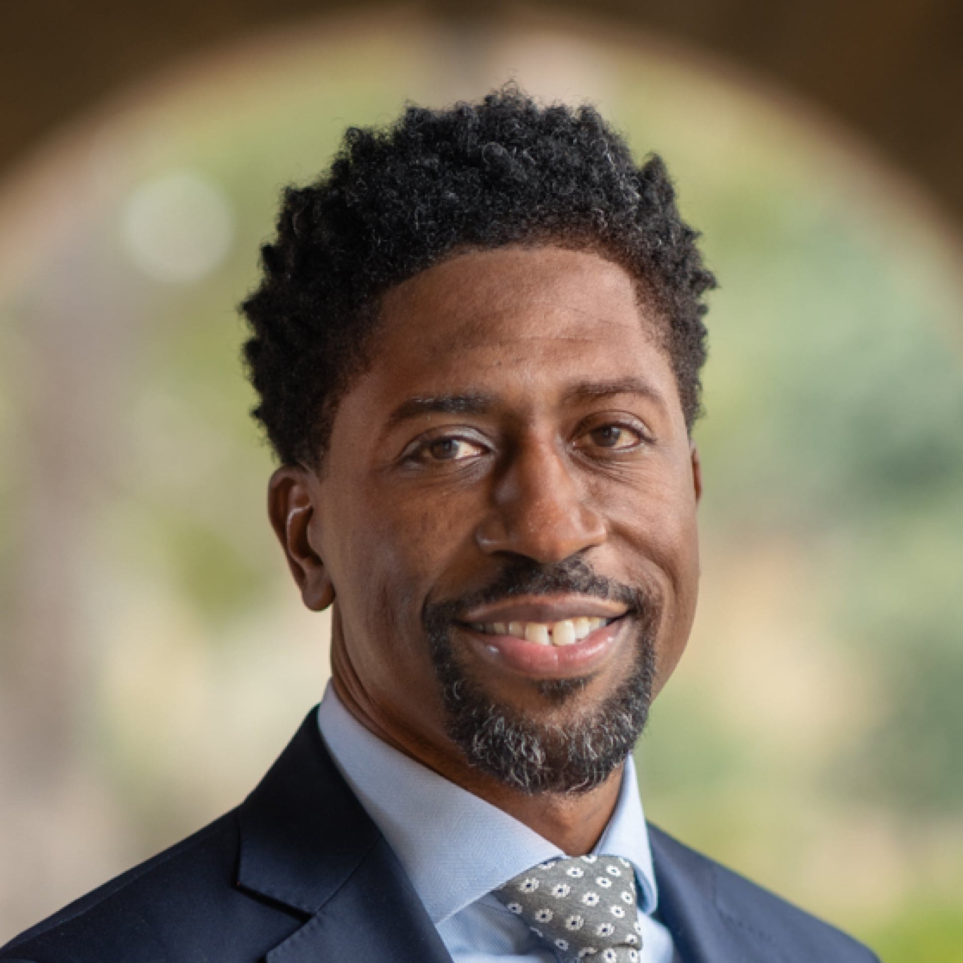 A man in a suit and tie smiles at the camera. He has short, curly black hair, a trimmed goatee, and stands in front of a softly blurred outdoor background.