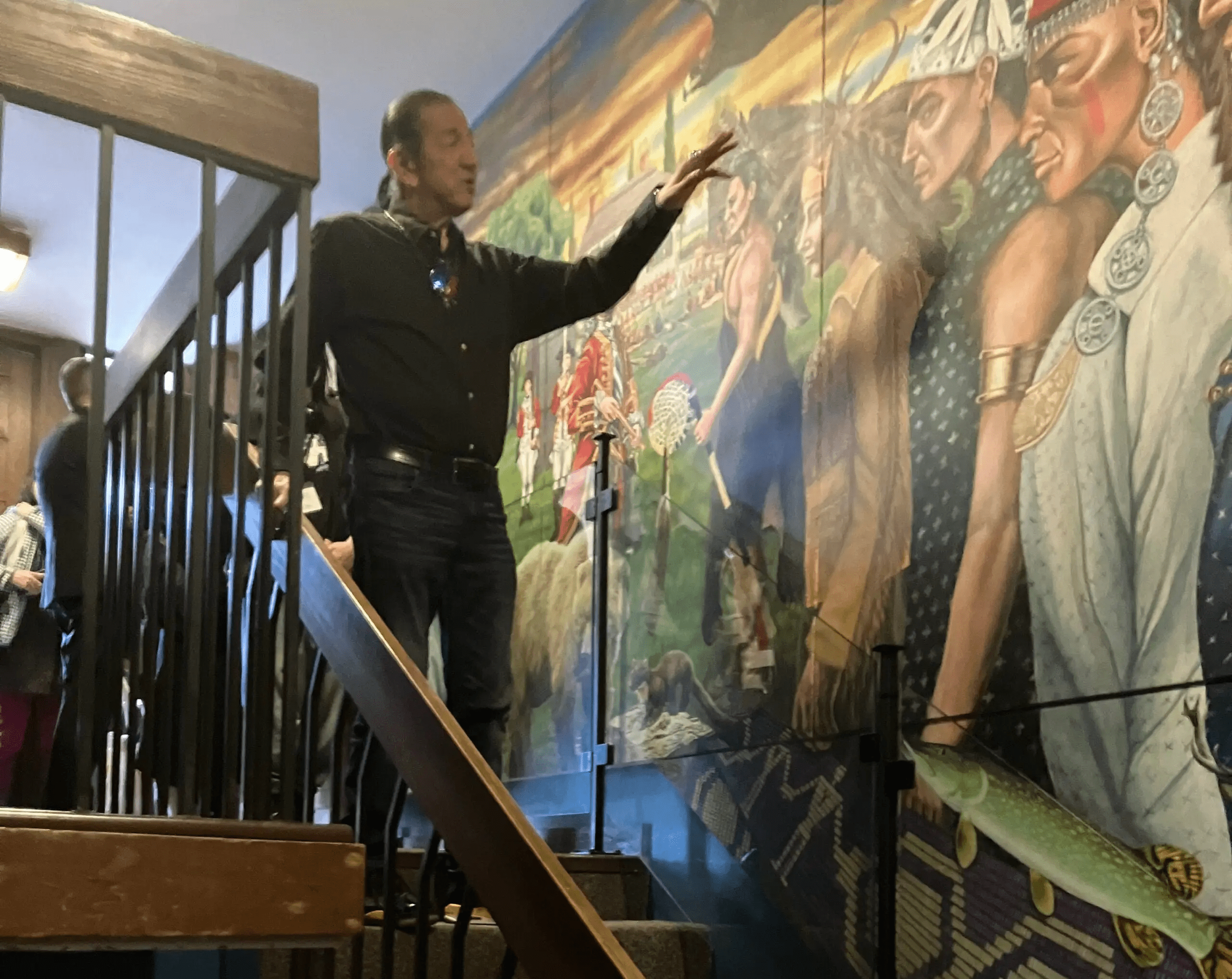 A man stands on a staircase, gesturing toward a vibrant mural depicting Indigenous people, traditional attire, and cultural scenes. Several people observe in the background.