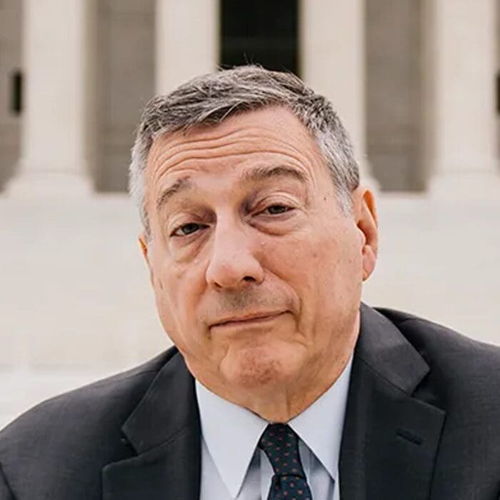 A middle-aged person with short gray hair, wearing a dark suit, white shirt, and dotted tie, stands outdoors in front of stone columns, looking directly at the camera with a neutral expression.