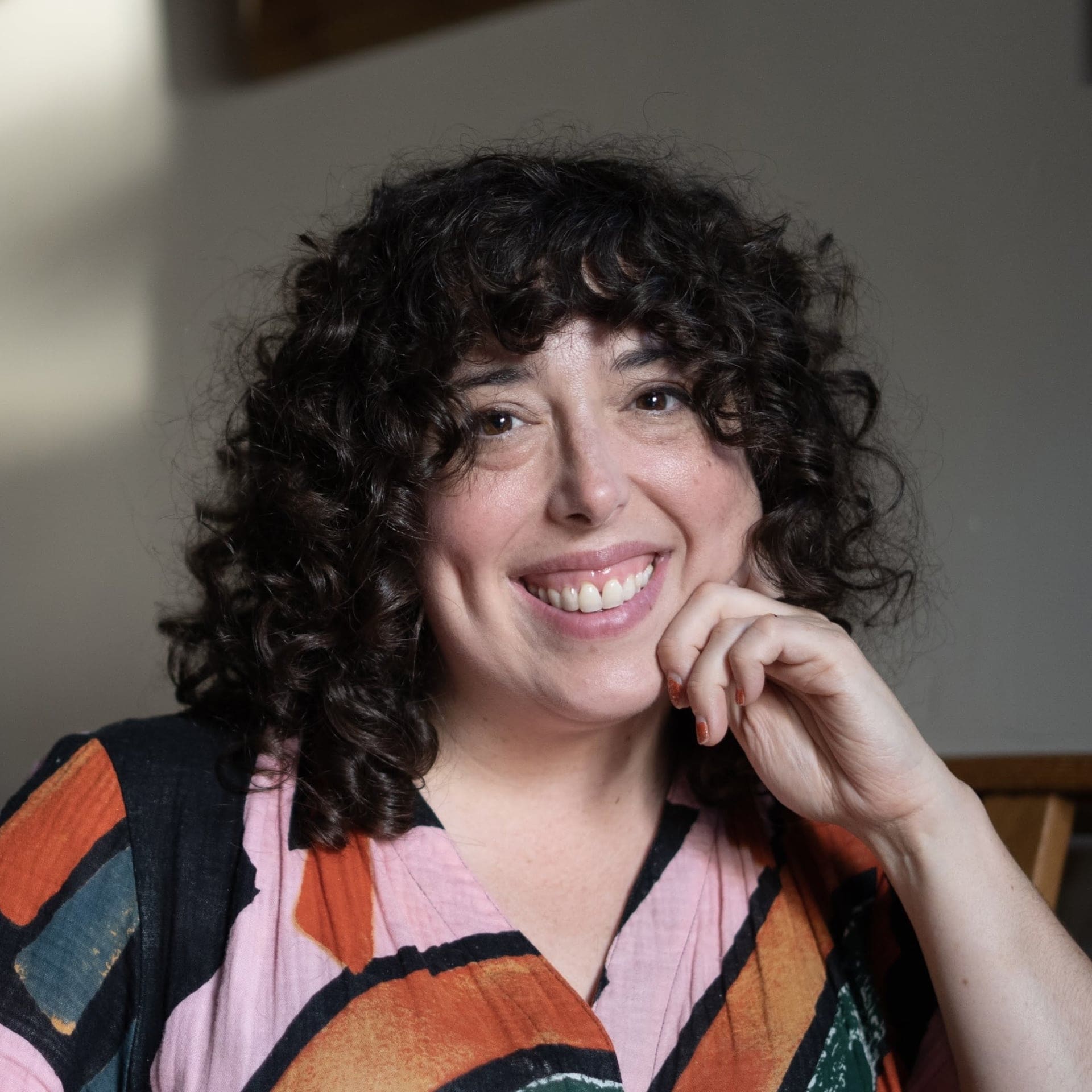 A woman with curly dark hair smiles warmly at the camera, resting her chin on her hand. She is wearing a colorful patterned top and is seated indoors, with soft light illuminating her face.