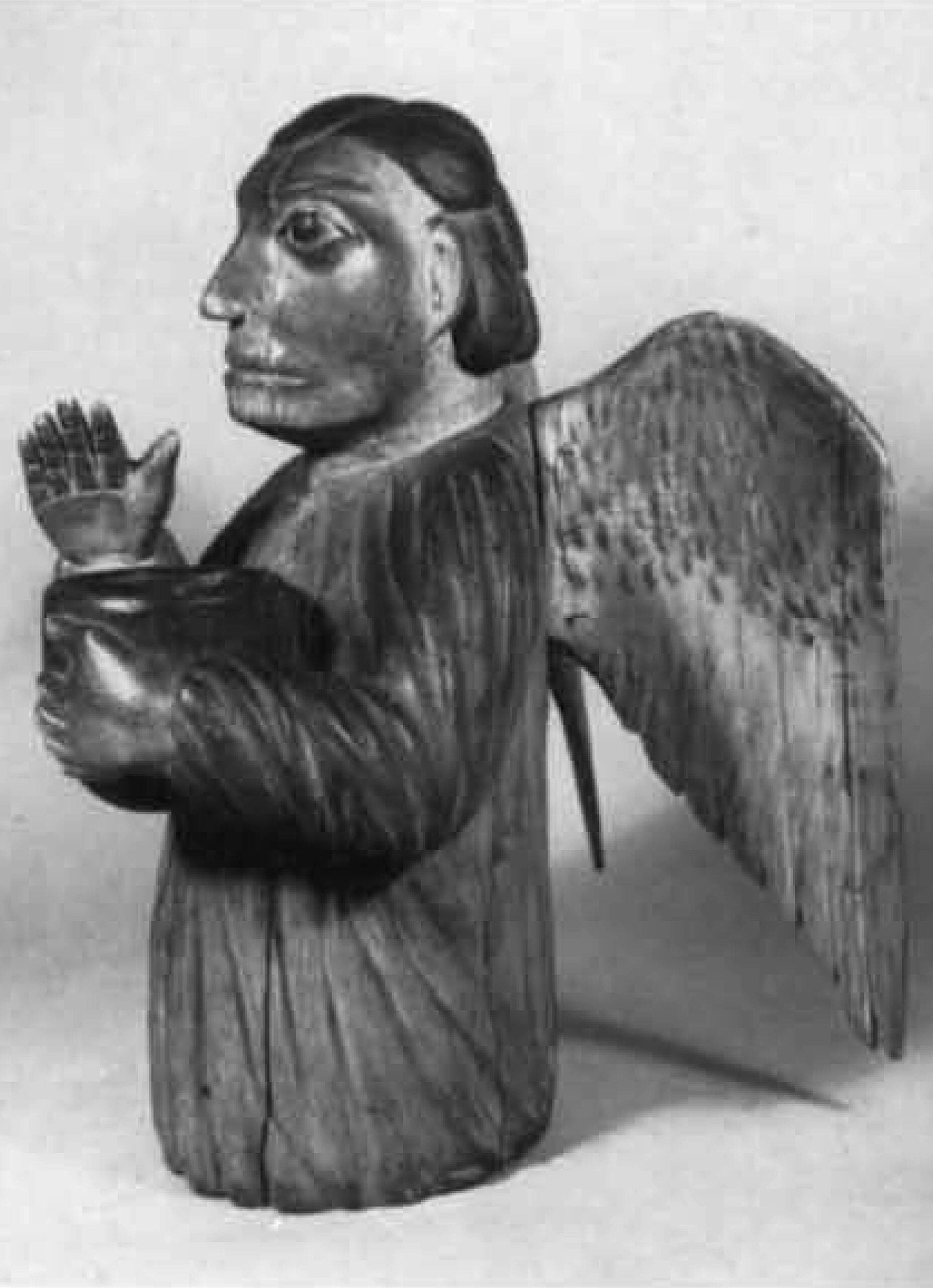 Wooden sculpture of an angel with large wings, raised right hand, and detailed facial features; the figure is dressed in a robe and has short hair, shown against a plain background.