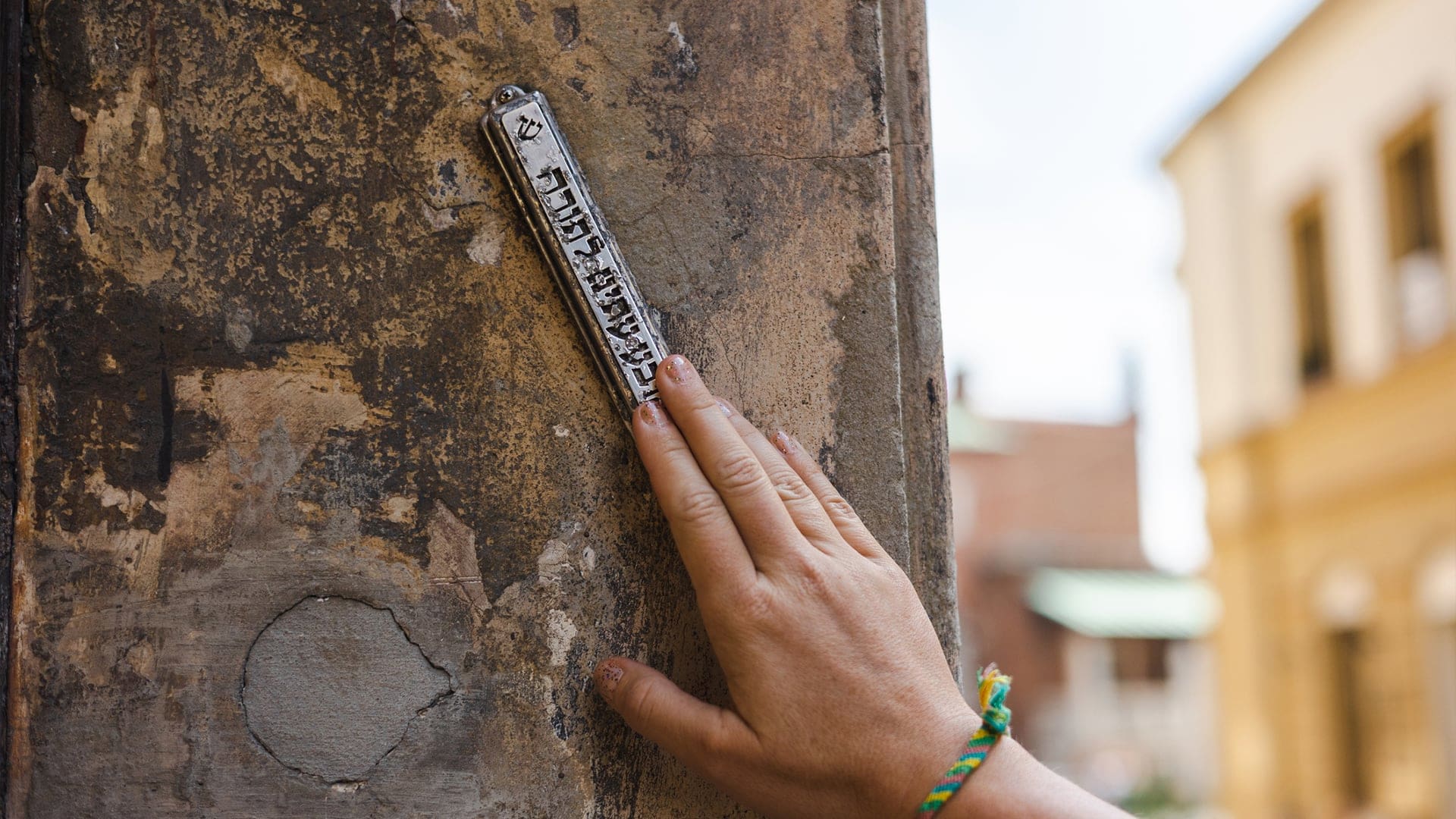A hand touches a mezuzah attached to a weathered doorpost, with Hebrew text visible on the mezuzah and blurred buildings in the background.