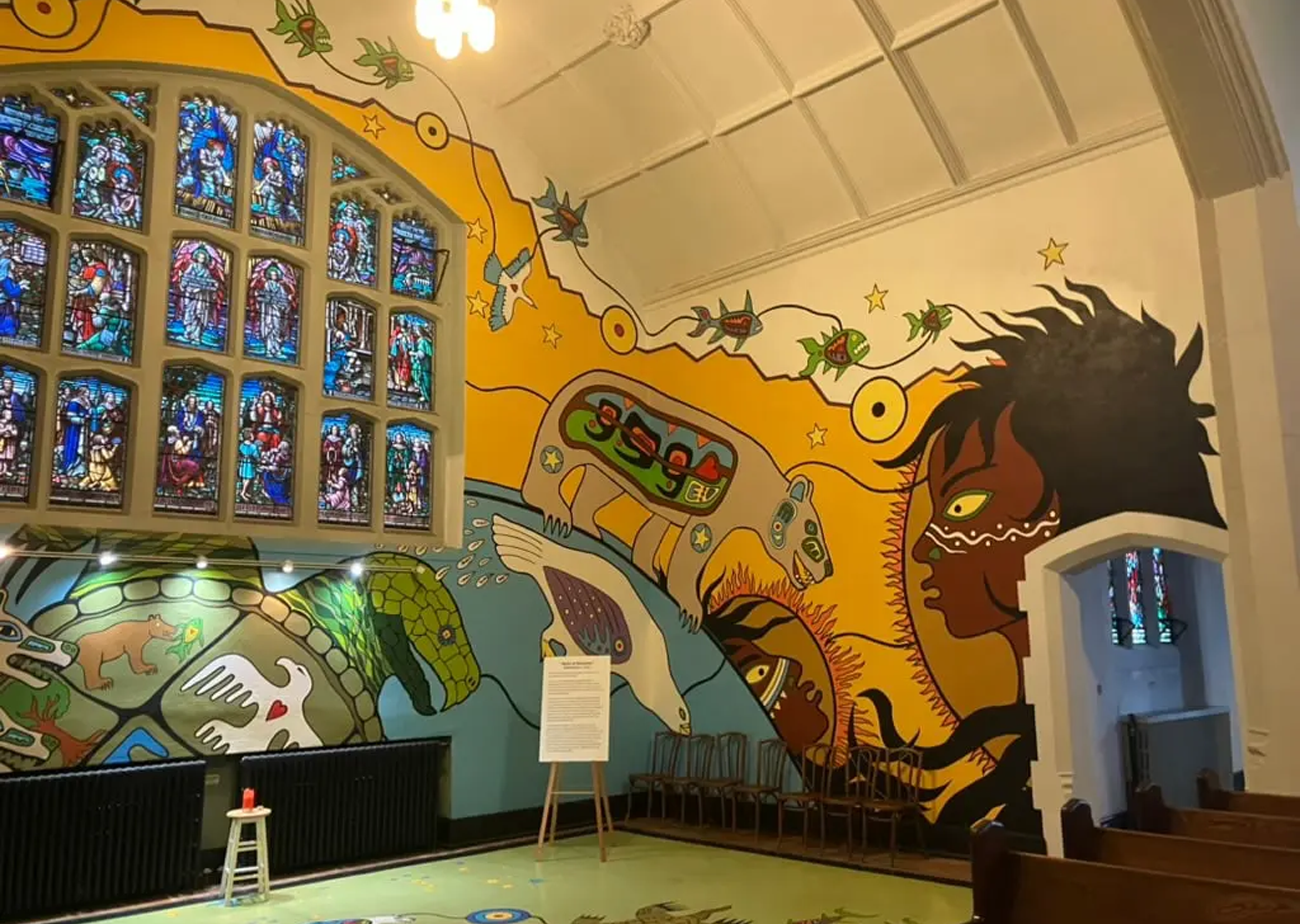 A brightly painted church interior features a colorful mural of animals, water, and a large human face, with stained glass windows on the left and wooden chairs along the wall. A sign and small red candle sit in the room.